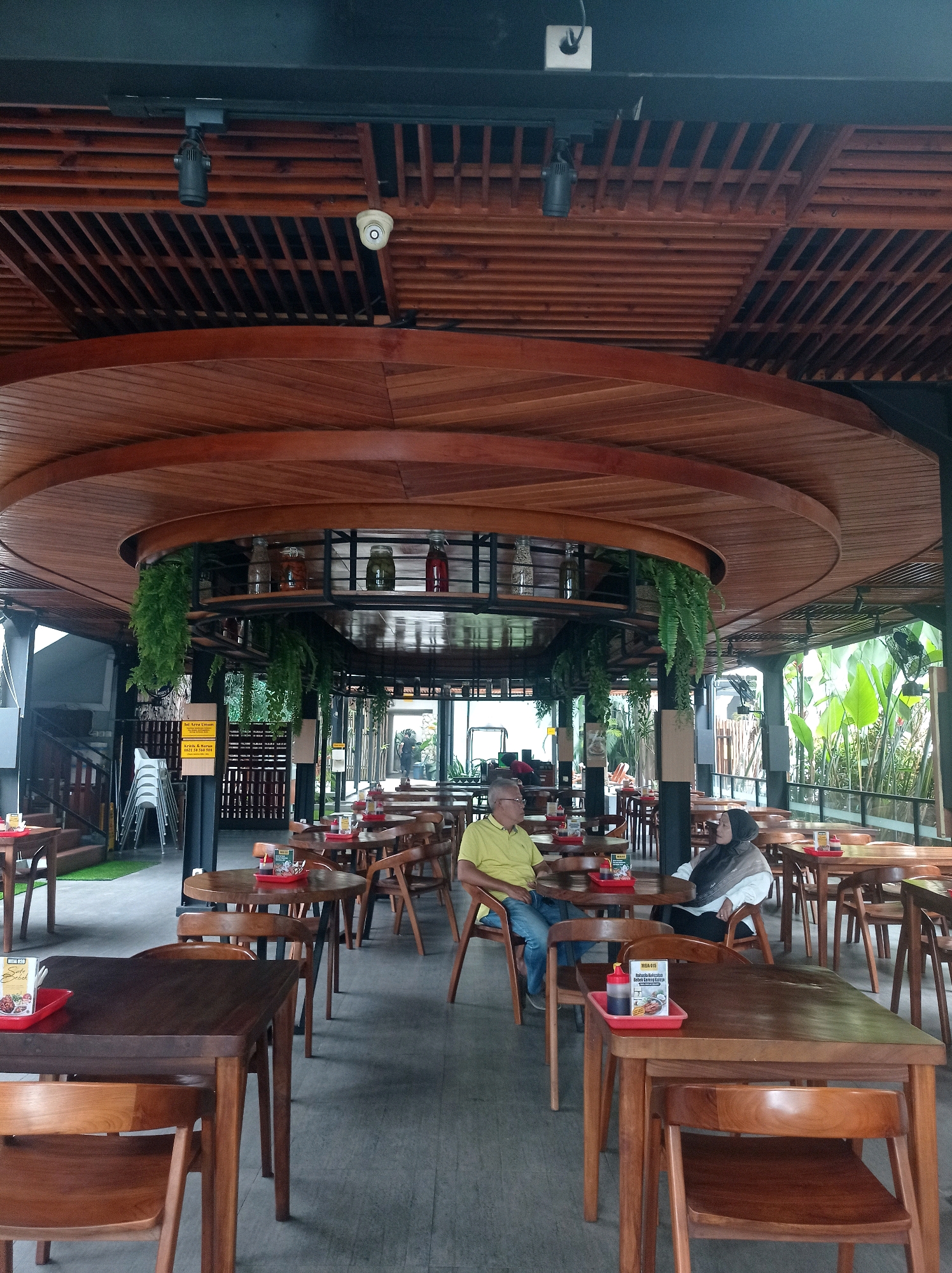 The image depicts the interior of a restaurant in Bandung City, Indonesia, characterized by its wooden architectural elements. A distinctive circular wooden ceiling structure dominates the upper portion of the scene, with wooden slats forming a decorative pattern. Suspended from this structure are several pendant lights and two CCTV cameras. Lush green ferns hang down from shelves, adding a touch of nature to the indoor setting.
The main area of the restaurant features numerous wooden tables and chairs arranged for dining. A couple is seated at a round table in the mid-ground, with a man wearing a yellow shirt and blue jeans, and a woman wearing a headscarf and a white top. Other tables in the foreground and background are unoccupied, some with red trays containing condiments. In the background, stacked chairs suggest a functional aspect of the establishment. Beyond the seating area, large glass doors open to an outdoor space with abundant greenery, indicating a connection to nature. The flooring is a light-colored, textured material. The overall ambiance suggests a modern, casual dining environment.