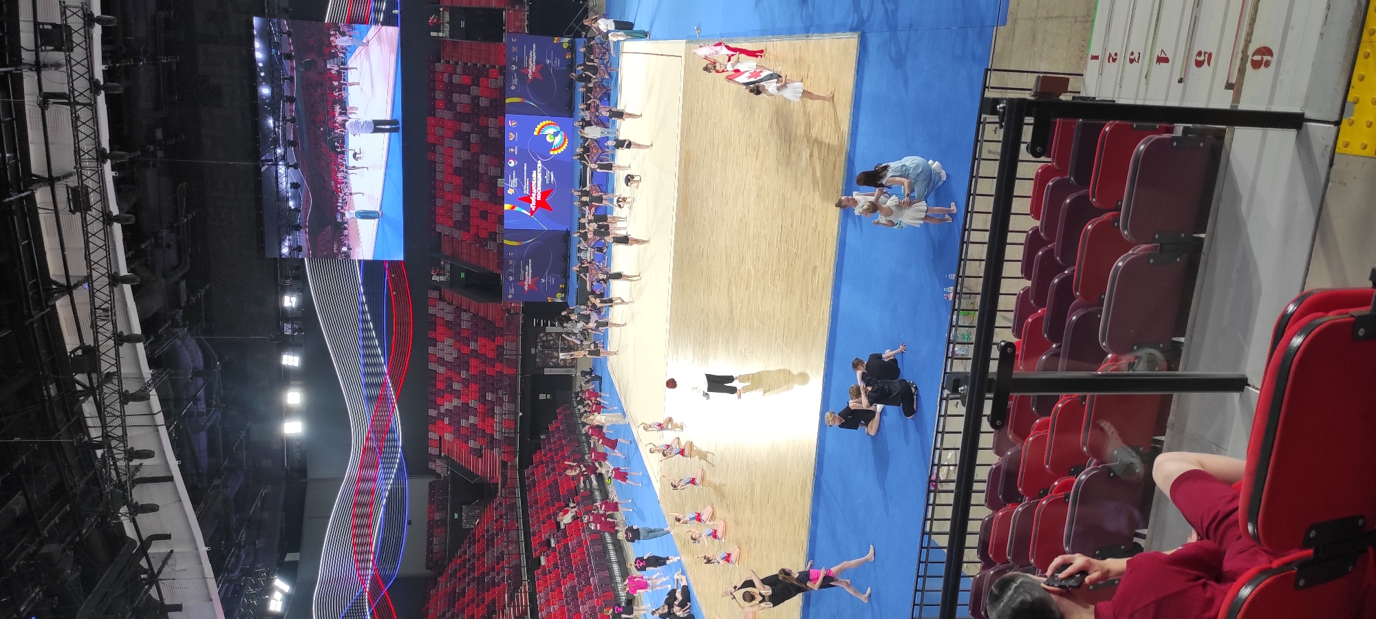 This image depicts a gymnastics event taking place in a large indoor arena, likely the Irina Viner-Usmanova Gymnastics Palace in Moscow, Russia. The arena features tiered seating, predominantly in red and dark red hues, surrounding a central wooden floor marked for gymnastics. A large video screen is suspended above the floor, displaying a live feed of the event.
On the floor, numerous young female gymnasts are engaged in various activities. Some are in formation, while others are practicing routines. Coaches or instructors, identifiable by their attire, are present on the floor, guiding the athletes. In the foreground, several individuals are seated in the stands, with one person in the lower left corner speaking on a mobile phone. The overall atmosphere suggests a competition or a large-scale training session.