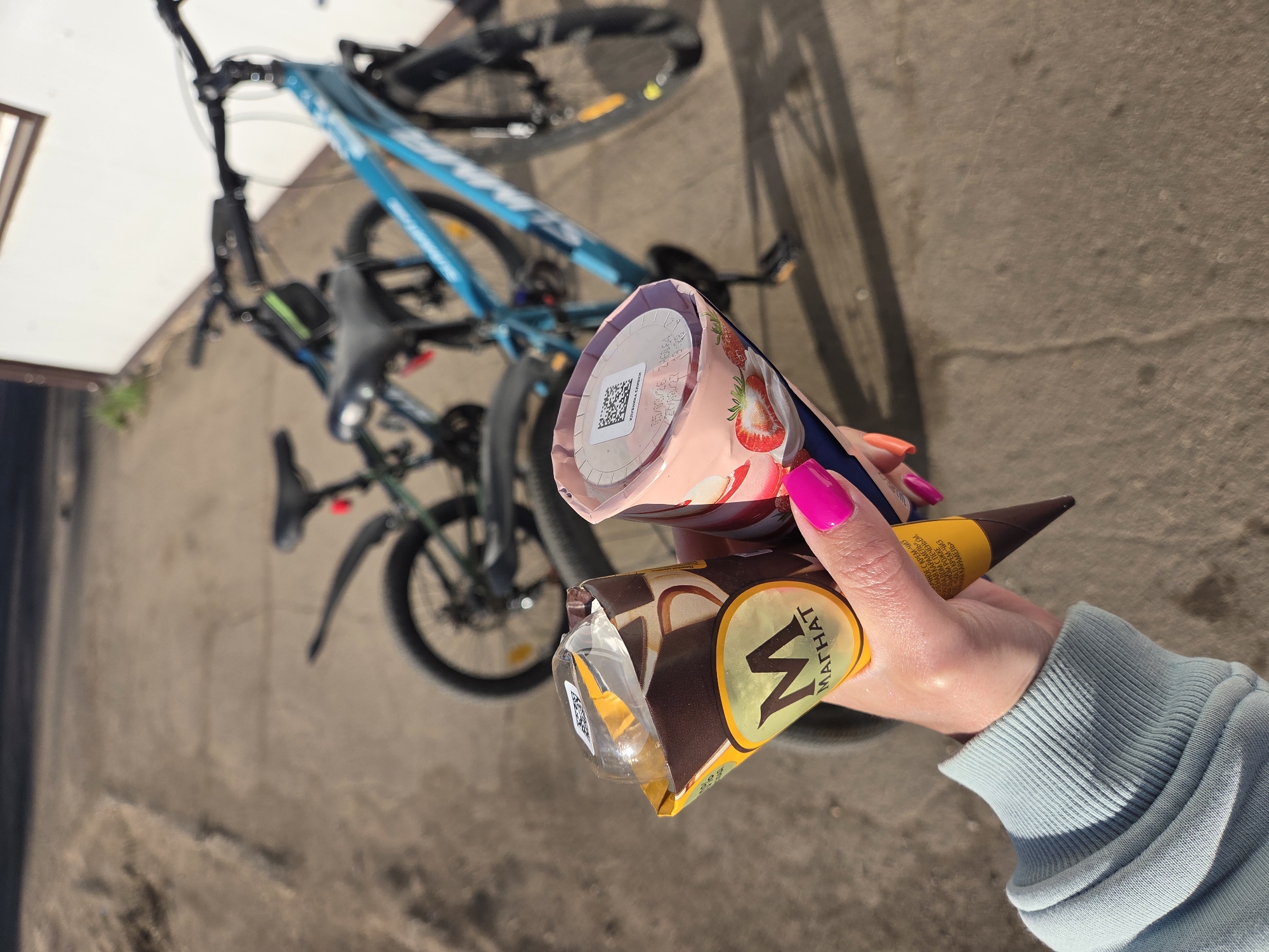 Here is a detailed description of the image:
The image presents a close-up view of a hand holding two ice cream cones against a backdrop of a bicycle and a paved surface. The hand, adorned with vibrant pink and orange nail polish, holds the ice creams with their branded packaging prominently displayed. One ice cream is a Magnum cone ("МАГНАТ" is visible on the packaging, which can be translated to "Magnum").
The background is somewhat blurred, but it is possible to identify a bicycle leaning against what appears to be a wall or building corner. The pavement has a weathered appearance.
The location seems to be Birobidzhan, Russia, based on the Cyrillic lettering on the ice cream packaging.