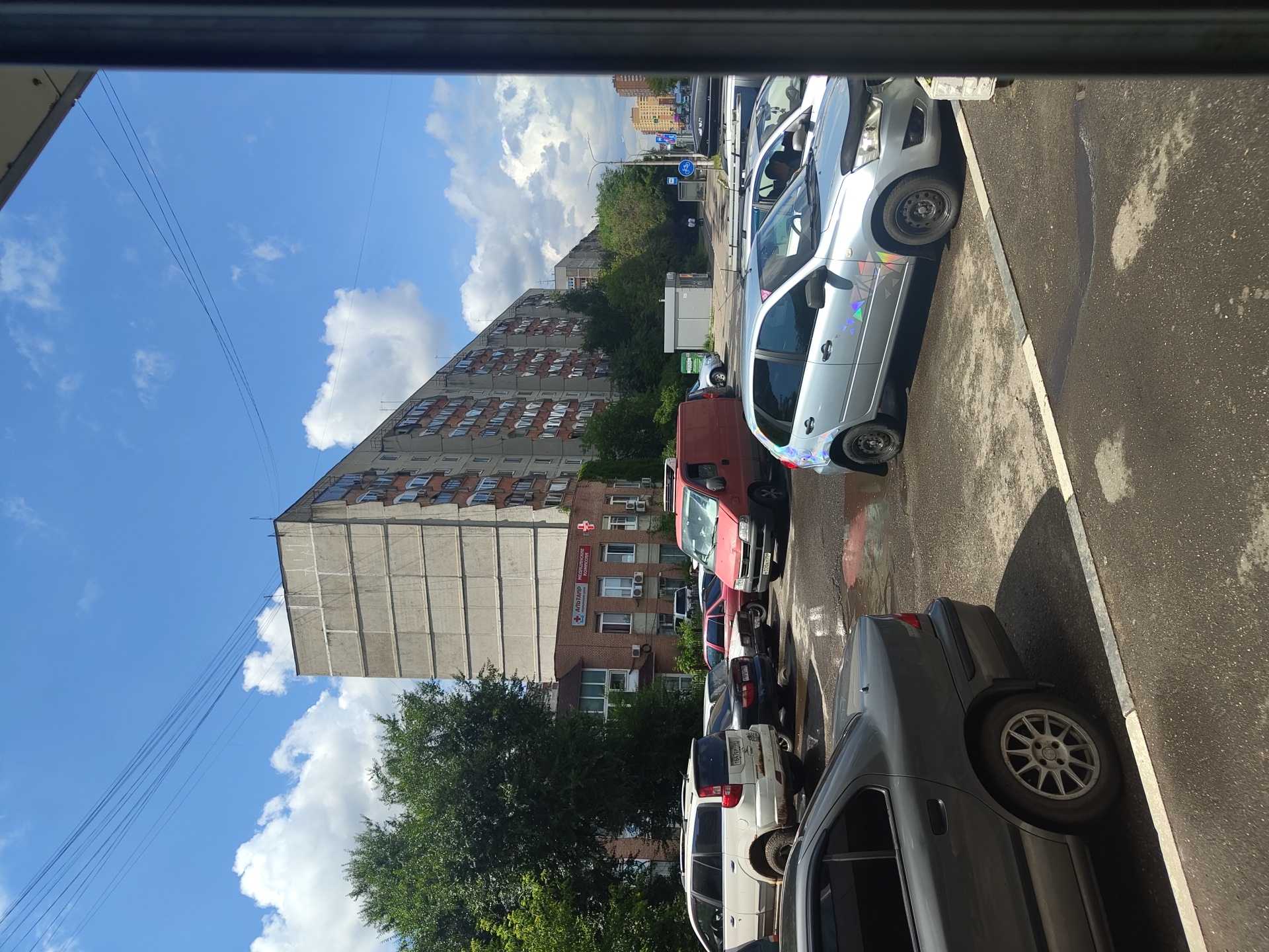 The image captures a street scene in Zhukovsky, Russia, on a sunny day with scattered clouds. In the foreground, a dark grey sedan is parked, facing away from the viewer. To its right, a silver hatchback is parked, its driver visible inside. Further back, a red van is parked, and beyond it, a white SUV is visible. A line of cars is parked along the side of the road.
Dominating the background is a tall, multi-story residential building with a concrete facade and balconies. To the left of the building, a smaller brick building is situated, bearing signage that reads "АЛЬТАР" and "МЕДИЦИНСКИЙ ЦЕНТР" (Medical Center) in Russian. Trees with lush green foliage line the street. The street surface appears wet in places, suggesting recent rain or cleaning. Overhead, power lines crisscross the sky. The overall impression is of a typical urban environment in a Russian city.