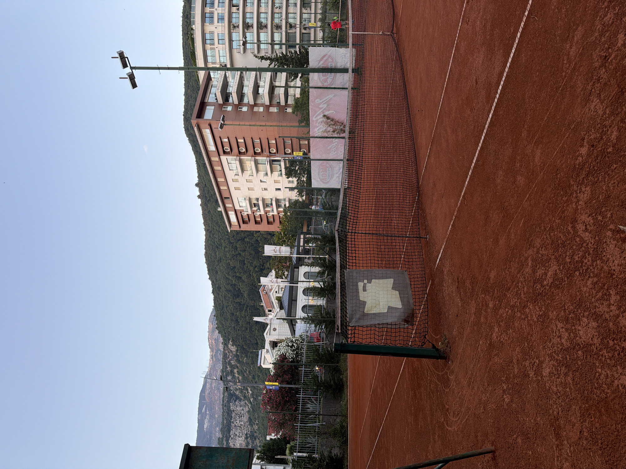 An outdoor clay tennis court is in the foreground of the image. A net divides the court in half. A sign hangs on the net. In the background, there are buildings, trees, and mountains. A single person, wearing a red shirt, is on the tennis court. The sky is clear and blue.