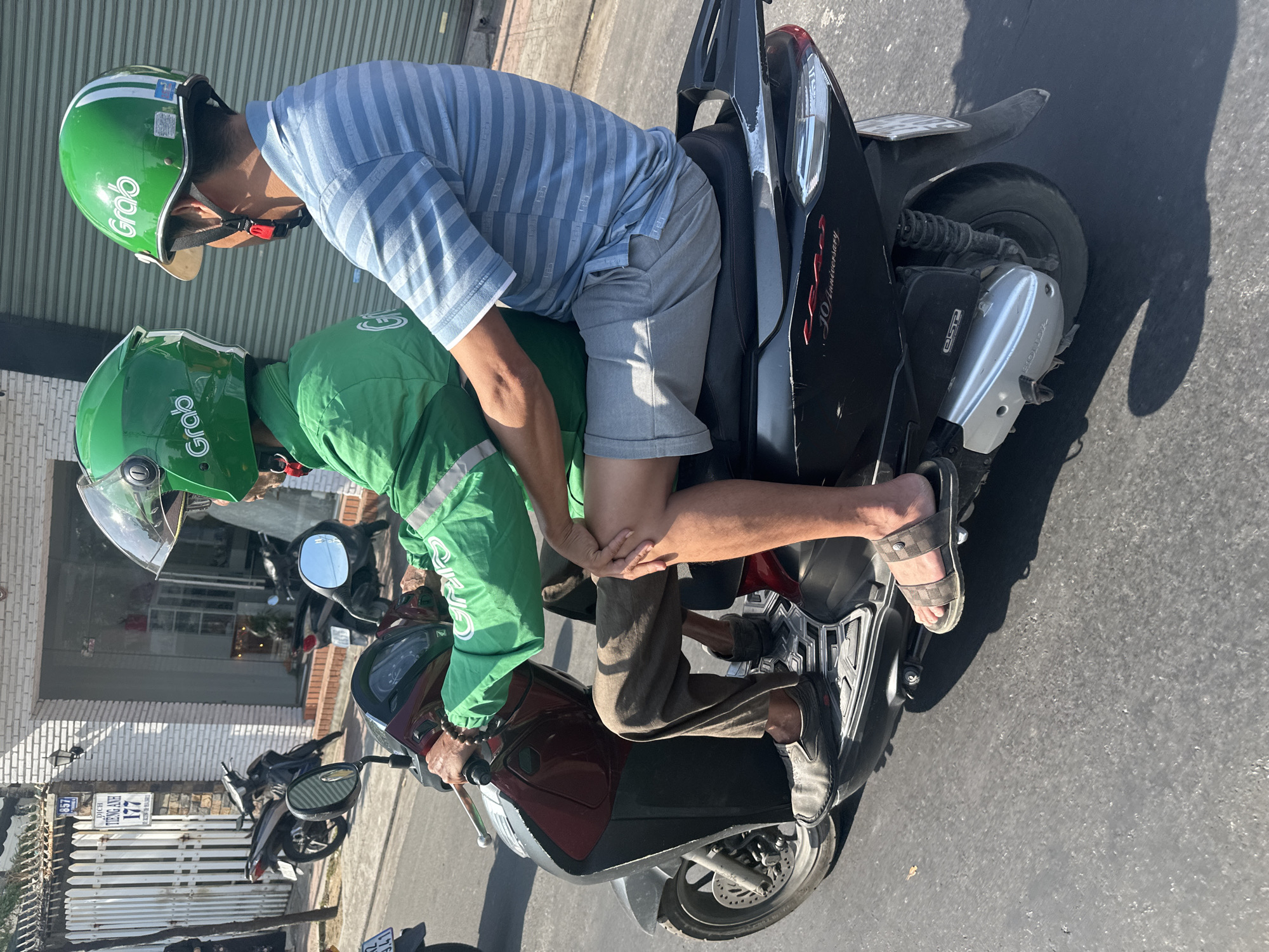 In Bac Nha Trang Ward, Vietnam, two individuals are riding on a black scooter. The person in front is wearing a green jacket with the "Grab" logo and a green helmet, while the person behind is wearing a blue and white striped shirt and grey shorts, also with a green "Grab" helmet. The scooter has a dark maroon front panel and black bodywork. The background features a white brick building with a metal fence and parked motorcycles, suggesting an urban environment. The road surface is asphalt.