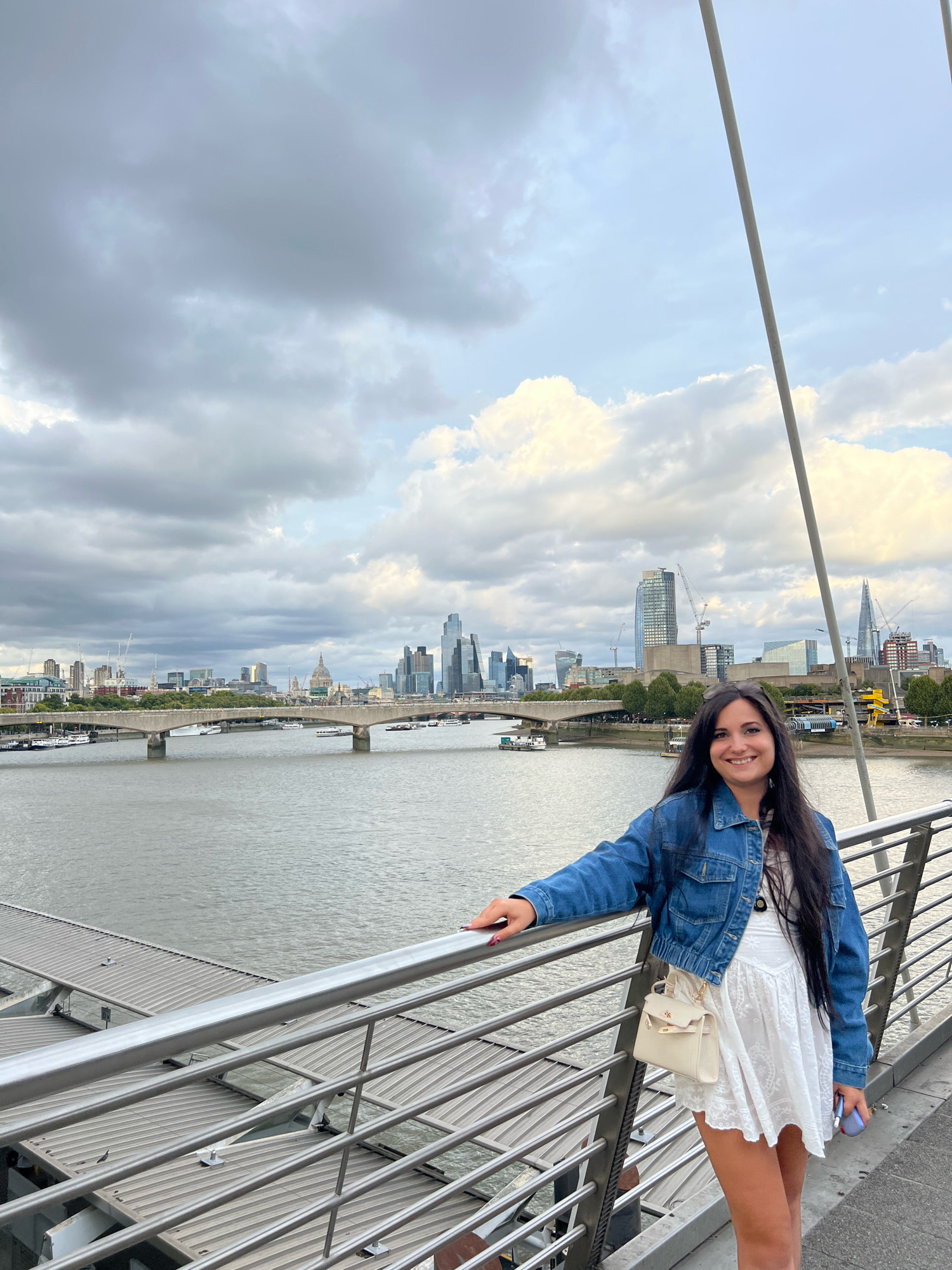 A full-body, slightly low-angle shot captures a smiling woman standing on a modern bridge overlooking the River Thames in London, United Kingdom. She is positioned on the right side of the frame, leaning her left arm on a silver metal railing. She has long, dark hair and is wearing a denim jacket over a white dress and carrying a small, cream-colored crossbody bag. Behind her, the river is calm, reflecting the overcast sky. In the background, a cityscape is visible, featuring the iconic landmarks of London, including various modern skyscrapers, a bridge spanning the river, and older buildings with domes. The sky is filled with dramatic, grey clouds, with patches of brighter sky peeking through. The bridge itself has a sleek, contemporary design with silver railings and cables extending upwards.