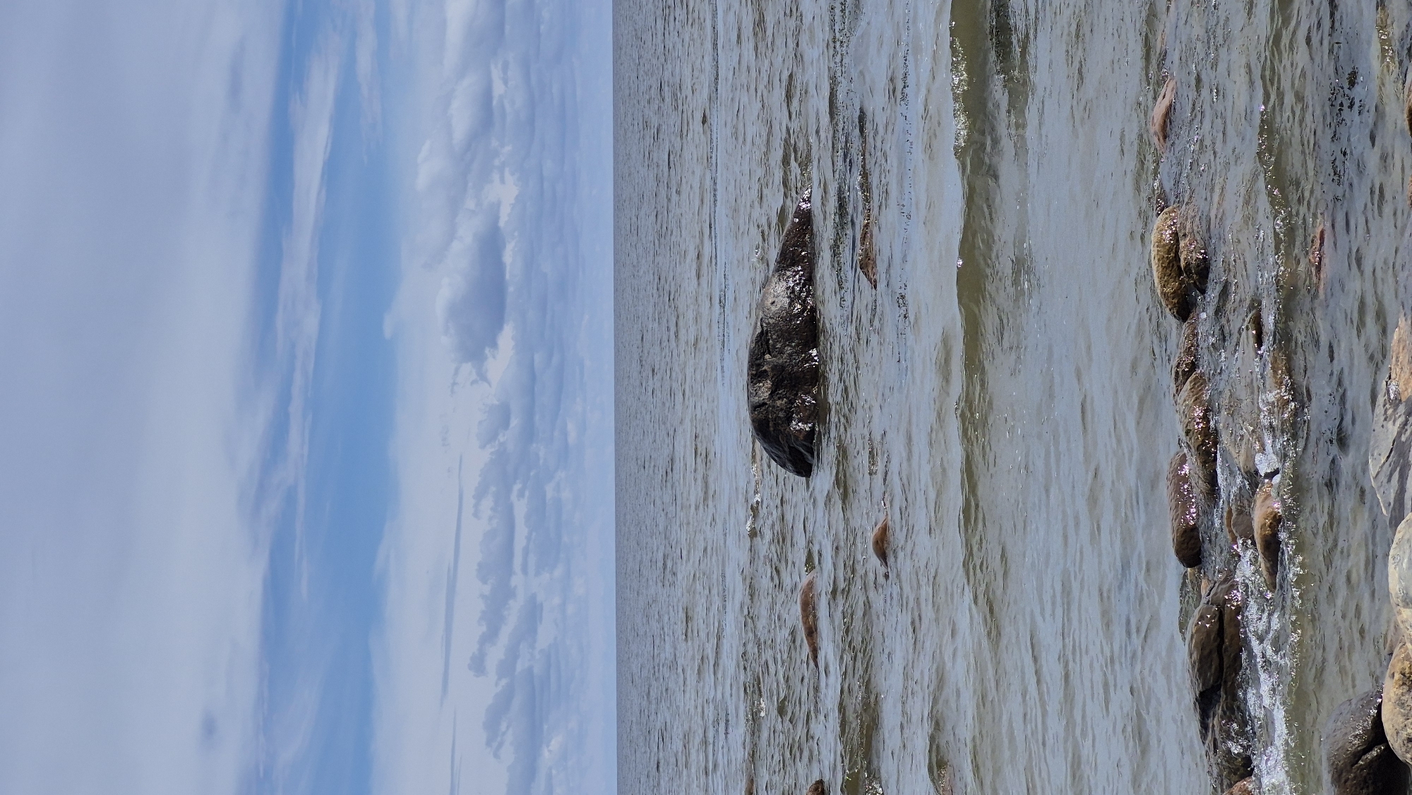 A calm sea under a cloudy sky is shown, with various rocks partially submerged in the water near the shore. The water is a muted gray-blue, with gentle ripples. The sky is a pale blue with scattered white and gray clouds. The rocks are dark brown and gray, with some showing a rough texture. In the foreground, a cluster of rocks breaks the surface of the water, creating small splashes. A larger, darker rock is prominent in the middle ground, with smaller rocks scattered around it. The horizon line where the sea meets the sky is clear and straight. The scene is tranquil and natural, with no human presence.