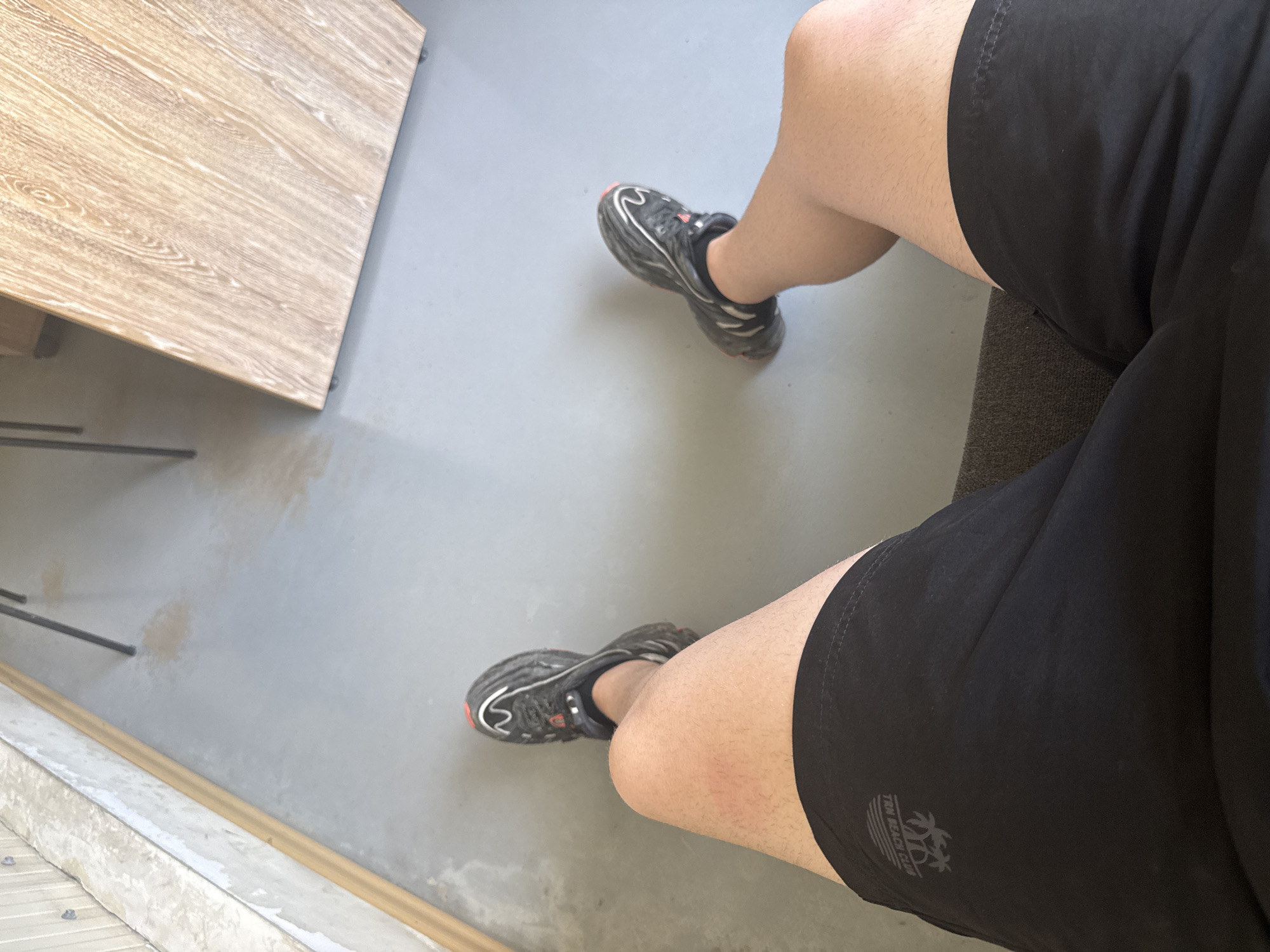 A first-person perspective shot shows a person sitting on a dark grey textured couch or bench. The person is wearing black athletic shorts with a light grey logo on the left thigh, and black and grey athletic shoes with red accents. Their bare legs are visible. The background consists of a light grey floor, a light-colored textured wall on the left, and a light brown wooden surface on the right, possibly a table or counter. The scene appears to be indoors. No specific landmarks of Stara Zagora, Bulgaria are visible in the image.