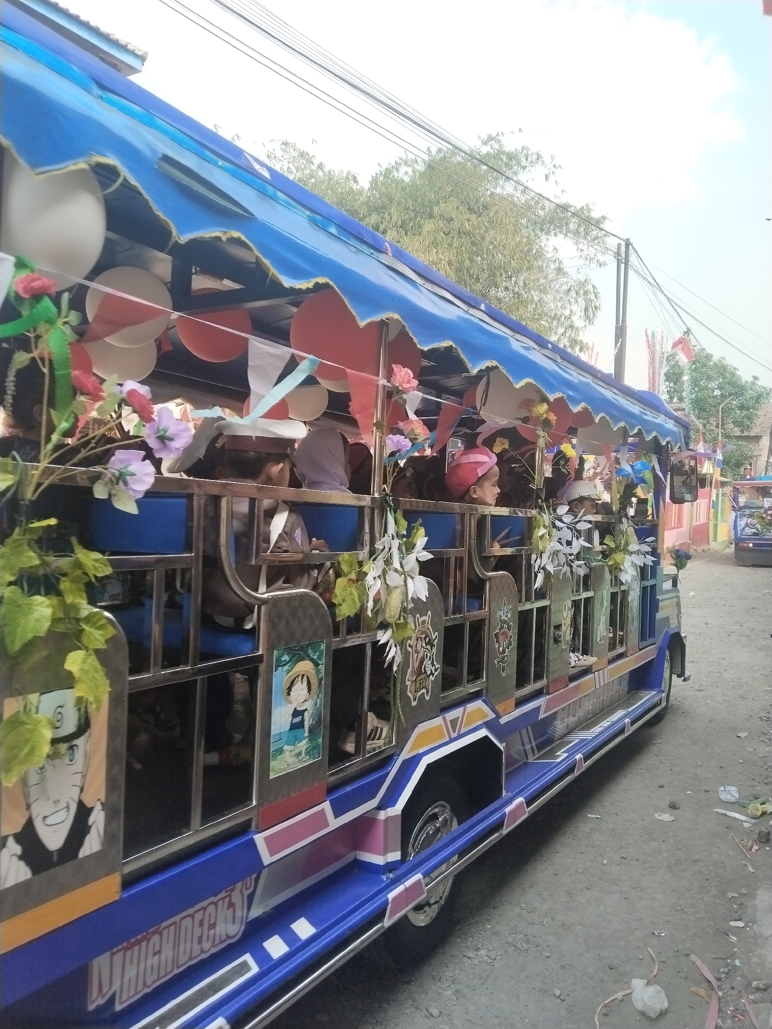 The image shows a brightly decorated jeepney, a common form of public transportation in the Philippines, parked on a dirt road. The jeepney is adorned with blue and red fabric canopy, balloons, and colorful flags. It also features images of anime characters on its sides. Several people, including children, are visible inside the jeepney. The background shows trees, utility poles, and houses, suggesting a residential or rural setting. The overall scene appears festive.