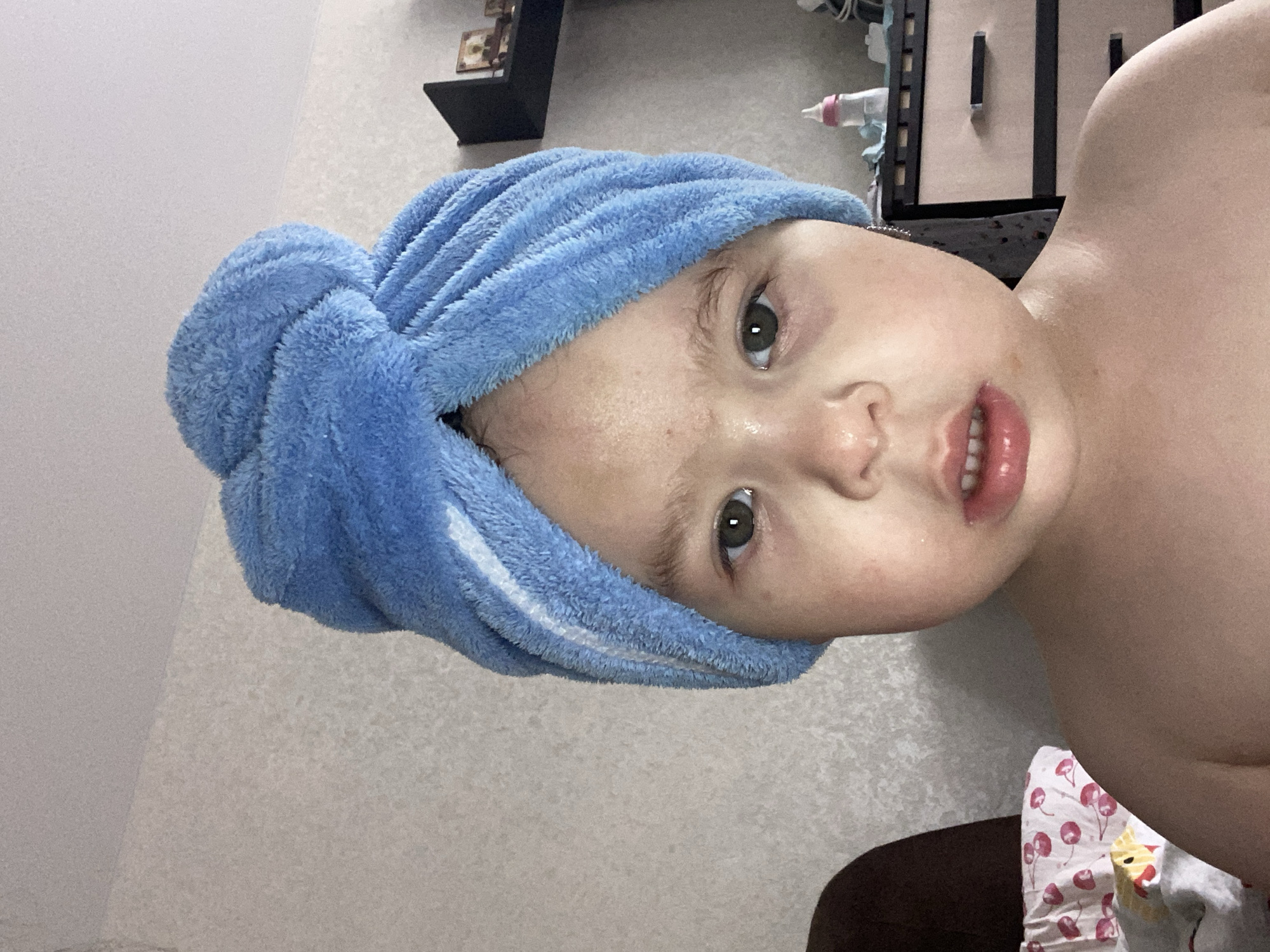 This media file features a close-up, rotated shot of a young child wearing a blue microfiber towel turban. The child has light skin, green eyes, and is looking towards the camera with an open mouth, showing teeth. Their cheeks appear flushed, and there are a few freckles visible on their face. The blue towel is wrapped around their head, with its textured fabric clearly visible.
In the background, a blurry scene includes what appears to be shelving with items on it, possibly in a room. There's also a glimpse of a baby bottle with a pink nipple. The lighting suggests an indoor setting.
The image focuses tightly on the child's face and upper body, emphasizing their facial expression and the towel. The overall impression is one of a candid, possibly post-bath moment. No specific time of day, weather, or explicit location cues are apparent, beyond it being indoors. There is no visible text in the image.