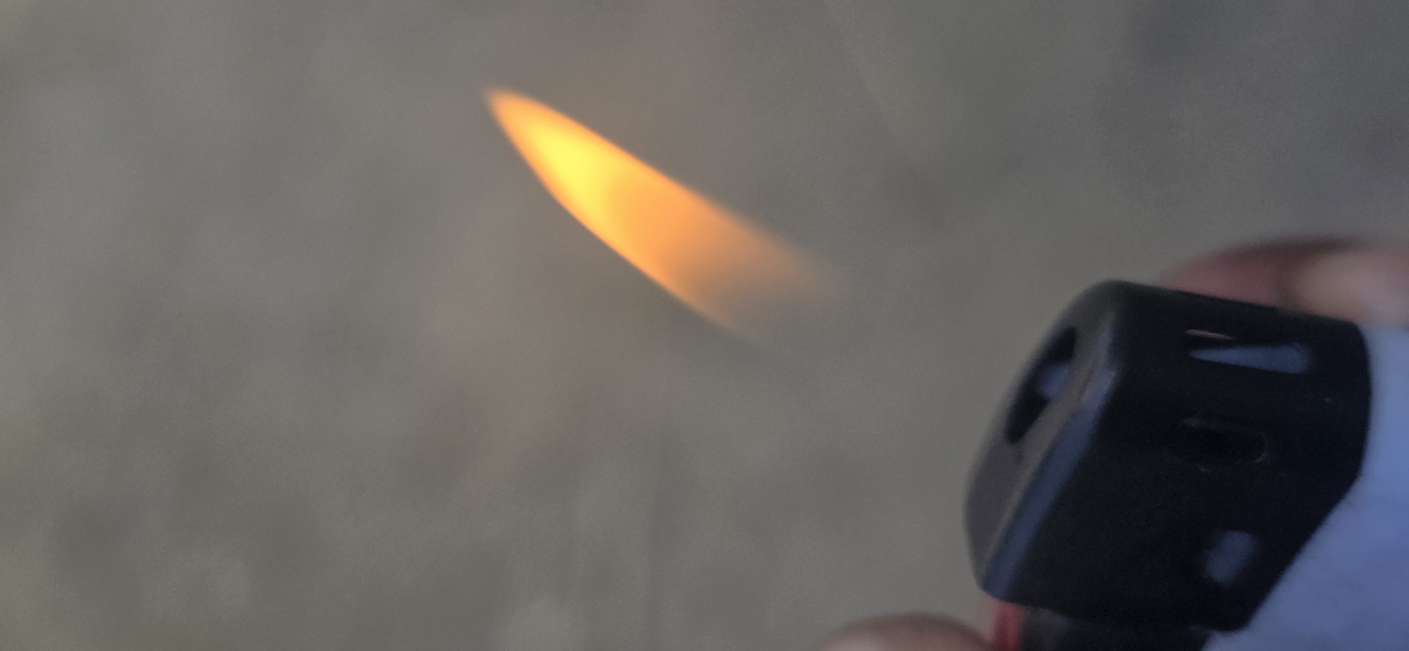 The image shows a close-up of a person's hand holding a black lighter with a flame. The flame is bright orange and yellow, tapering to a point. The background is out of focus, appearing as a soft, muted gray. It is not possible to determine the time of day, weather, or specific location from the image, beyond the provided context of Nizhnevartovsk, Russia. No text is visible in the image. The image captures a simple, everyday action of igniting a flame.