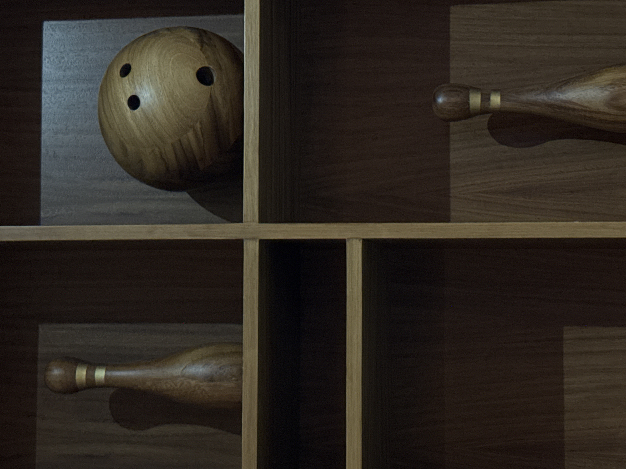 The image shows a wooden shelf with a bowling ball in the upper left compartment and two bowling pins, one in the upper right and one in the lower left compartment. The scene is dimly lit, with shadows indicating a light source from the upper right. The wood has a visible grain pattern. No people are visible. The location is not possible to determine from the visual information provided.