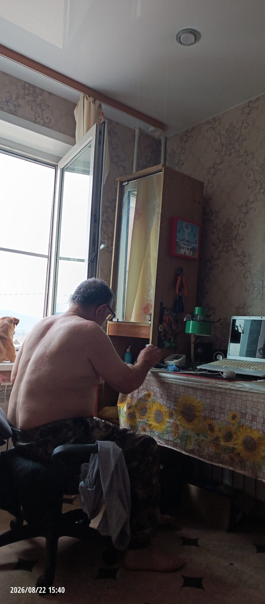 A bare-chested man with glasses sits at a table in what appears to be a room with patterned wallpaper and a white ceiling. He is facing away from the camera and is positioned to the left of the frame, his back dominating the left half of the image. He appears to be engaged in an activity at the table, with his hands in front of him.
The table is covered with a checkered tablecloth featuring a sunflower print. On the table, to the right of the man, is a laptop, a green desk lamp, and various other objects, including what appears to be a blue bottle.
To the left of the man, a window provides a view of an outdoor scene, with what looks like mountains in the distance and a sliver of a sky. There is also a light-colored dog visible on a windowsill.
Further to the right in the image, there is a tall wooden cabinet with a mirror on its door. A red clock hangs on the cabinet, and several items, possibly tools, are suspended below it.
The floor is tiled with a dark pattern. A black office chair is partially visible in the foreground to the left, with clothing draped over its armrest. The overall impression is of a domestic interior, possibly a home office or living space. The timestamp in the bottom left corner indicates the image was taken on August 22, 2026, at 15:40.