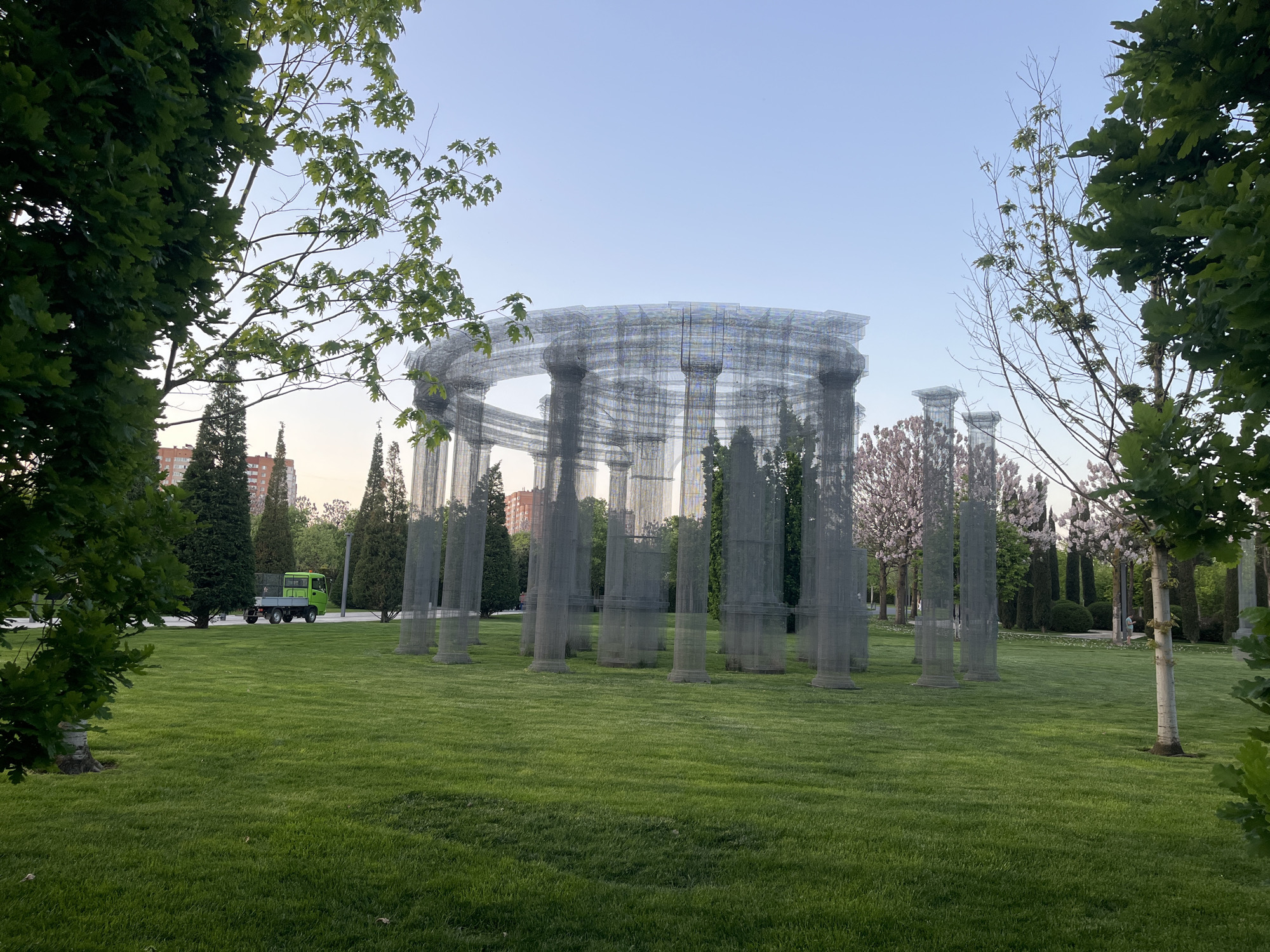 The image depicts an art installation resembling a classical rotunda composed of wire mesh, situated on a green lawn in a park. The rotunda is encircled by trees of varying species, with some in bloom. In the background, evergreen trees and a building are visible. A small green utility vehicle is parked near the trees to the left of the rotunda. The sky is a pale blue, suggesting early morning or late afternoon light. The location is believed to be in the city of Krasnodar, Russia. There are no people clearly visible interacting with the scene.