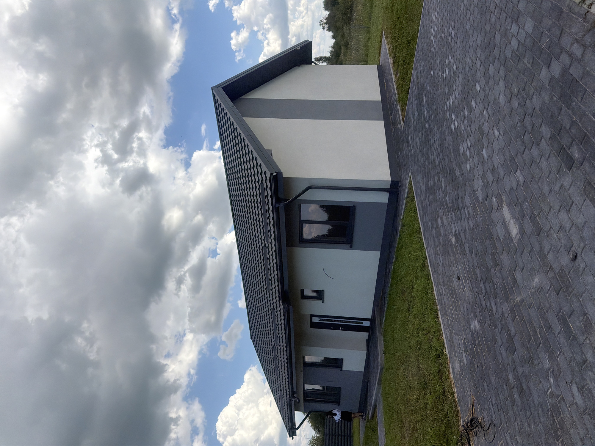 A young woman stands on a paved pathway in front of a modern, single-story house in Golubevo, Russia. The house has a dark gray tiled roof, white and gray exterior walls, and dark-framed windows. A black-and-white tiled pathway leads up to the entrance. The surrounding area is covered in green grass, and a dense, green hedge is visible on the left side of the image. The sky is filled with dramatic clouds, with patches of blue visible.