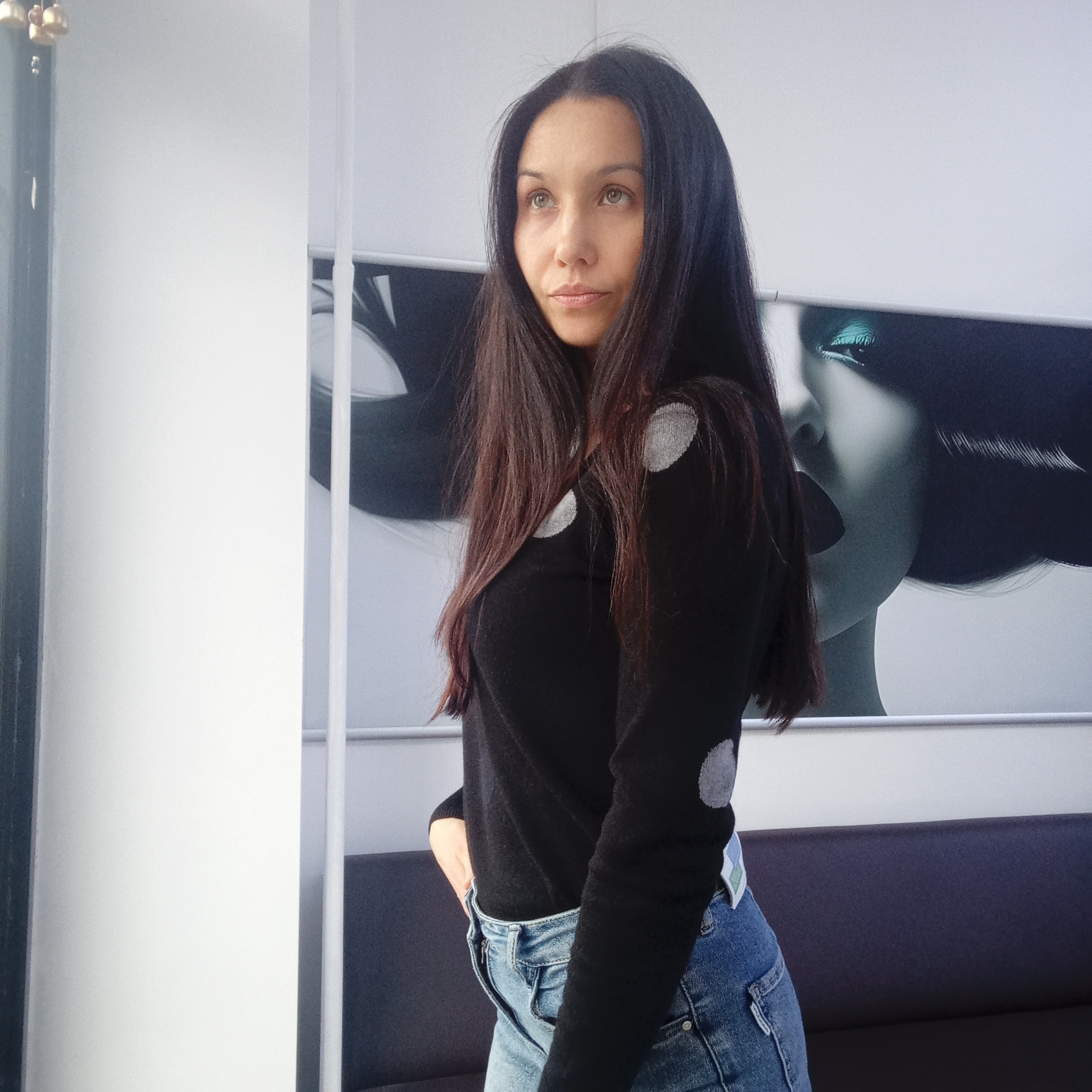A woman with long, dark hair stands indoors, looking slightly upwards and to the left. She is wearing a black, long-sleeved sweater with grey polka dots and blue jeans. She has one hand tucked into her jeans pocket. Behind her is a wall with a black and white print of a woman's face, featuring turquoise eyeshadow. A dark grey couch sits beneath the print. To the left of the woman is a white wall with a vertical support column, and a dark window frame is slightly visible in the very left corner. The location is most likely Izhevsk, Russia, based on the image context.