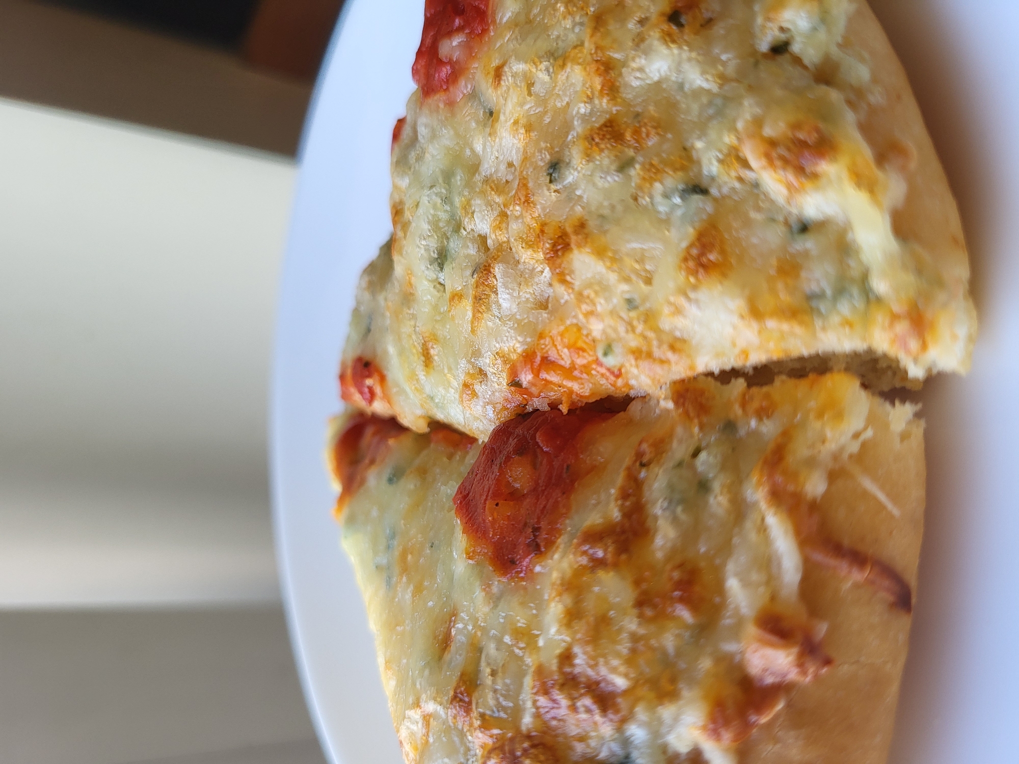 Two slices of pizza are on a white plate, close up. The pizza has a golden crust, topped with melted cheese and tomato sauce. The lighting suggests the pizza is near a window, with soft light illuminating the scene. The background is out of focus, showing neutral-colored curtains or blinds and a glimpse of a dark object that could be furniture.