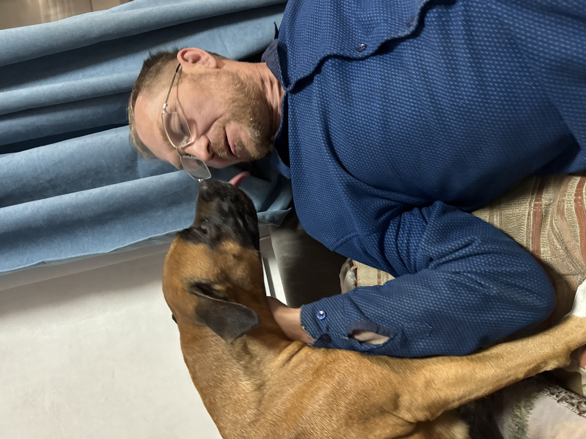 A middle-aged Caucasian man wearing glasses and a blue textured shirt is lying down, face to face with a brown and black dog. The dog is licking the man's face. A light blue, textured blanket is visible in the background. The location appears to be indoors. There is no evidence to suggest that this picture was taken in the city of Chaikovsky, Russia.
