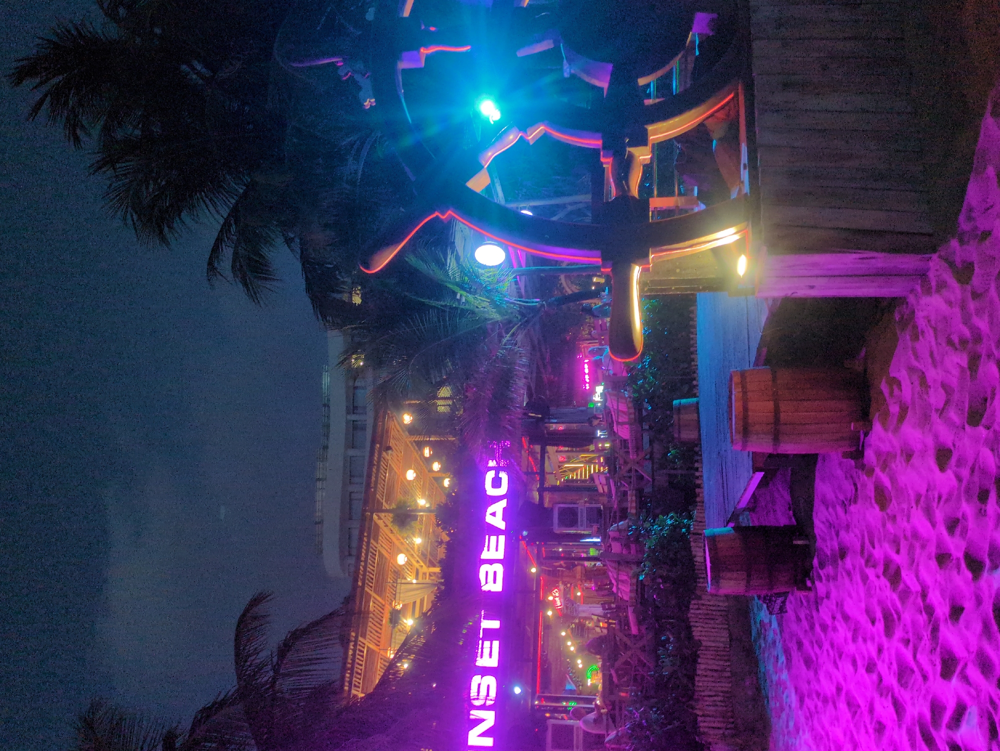 At night, a photograph of “Sunset Beach” in Phú Quốc, Vietnam, taken at an angle, shows the illuminated beachfront area. The scene is anchored by a palm tree in the left foreground against a dark, clouded sky. The “SUNSET BEACH” sign is vertically oriented and brightly lit in purple neon, drawing the eye toward a multi-level, open-air bar or restaurant structure. The wooden building is adorned with string lights. The right side of the frame is a ground-level area, perhaps sand, with wooden barrels or stools, lit in an intense purple hue. Overhead, an artistic installation with wooden elements and vibrant blue and red neon lights adds a modern touch. No people are visible.