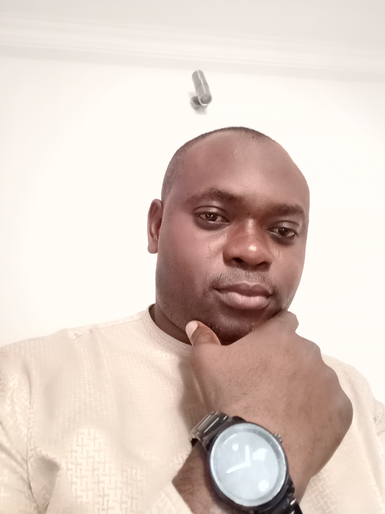A close-up, eye-level shot shows a dark-skinned man indoors, likely in Gwarinpa, Nigeria. He is balding, wearing a light-colored, patterned shirt, and a wristwatch with a dark band. His hand is raised to his chin in a contemplative gesture. The background is a plain, light-colored wall with a silver, cylindrical object affixed to it near the top of the frame. The lighting appears artificial. No other people or objects are visible.