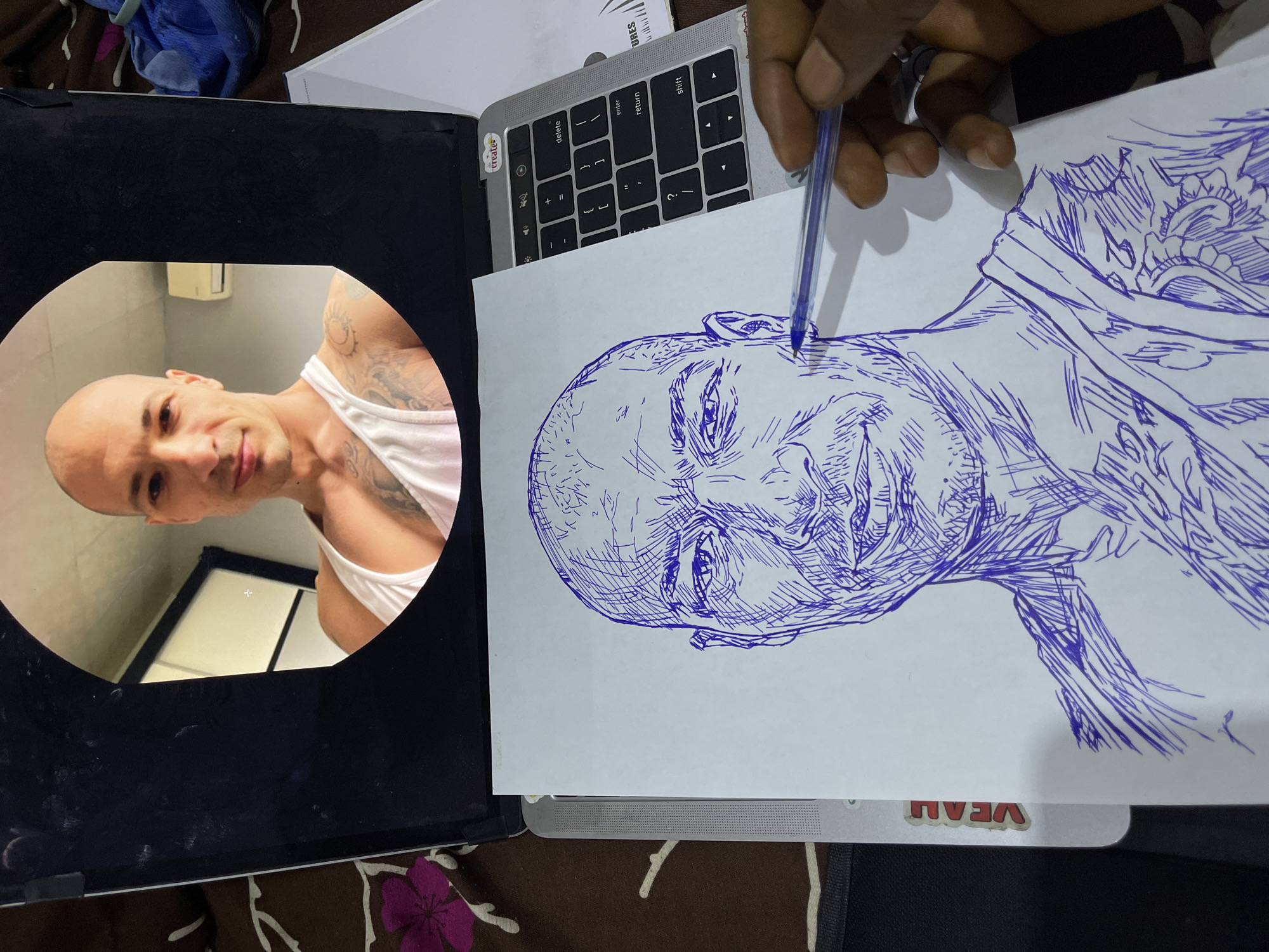 The image shows a man's face being drawn on a piece of white paper with a blue pen. The drawing is a sketch of a bald man with tattoos on his shoulder and chest. The paper is placed on a laptop keyboard. The laptop screen displays a circular image of the man who is being drawn. The background shows a cluttered desk with various items, including a blue towel and a notebook. The image appears to be taken from a top-down perspective, with the hand holding the pen visible in the bottom right corner. There is no indication of the location in Abuja, Nigeria.
