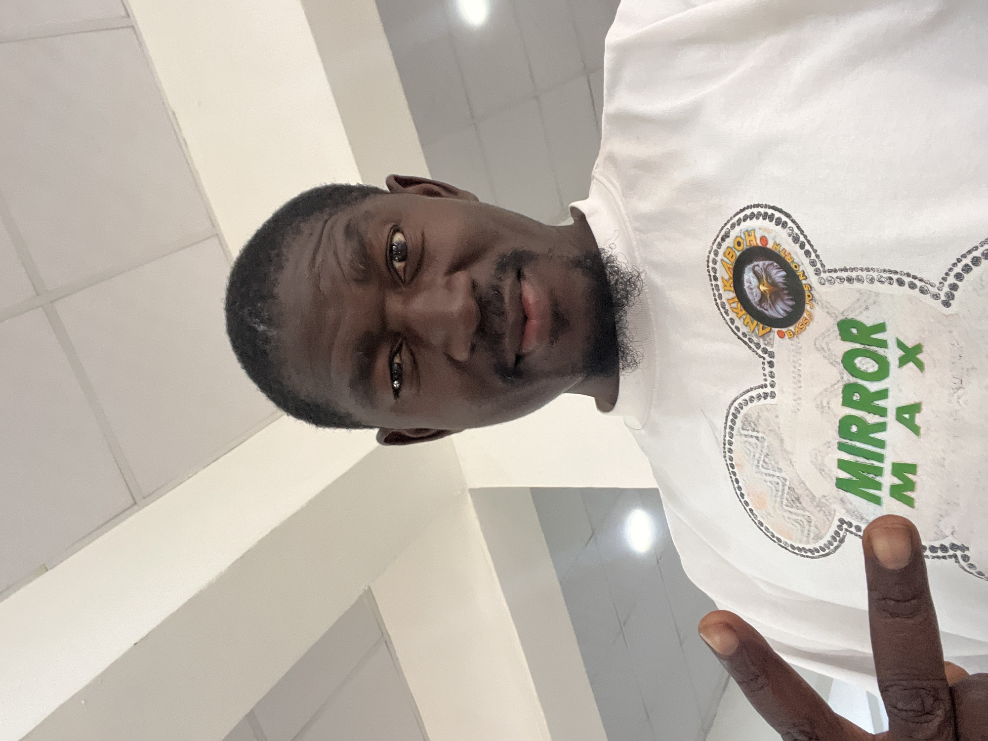 A man is taking a selfie in what appears to be an indoor setting with a white ceiling and fluorescent lighting. He is wearing a white t-shirt with a graphic design that includes an eagle and the text "MIRROR MAX" and "ANKI KABO BASSA JOS NORTH". He is making a peace sign with his left hand. The image quality is moderate, with some pixelation noticeable.