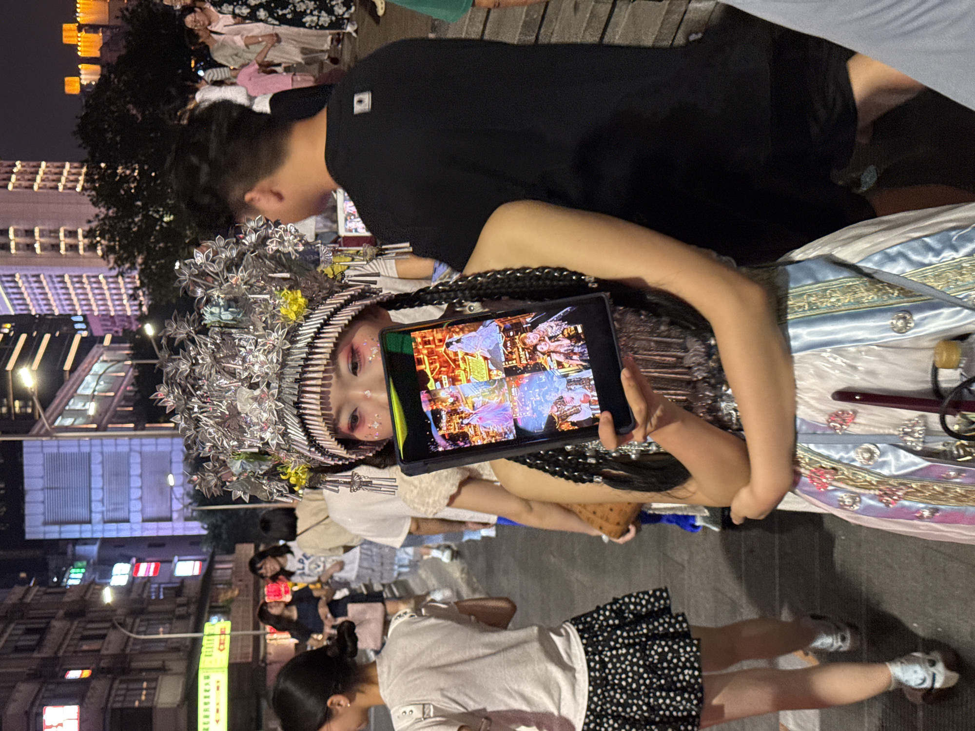 In the city of Yuzhong District, China, at night, a young woman adorned in a traditional, elaborate headdress and makeup holds up a tablet displaying four images. She wears a white and blue ornate dress with ribbons and embellishments. The tablet shows images of traditional Chinese cultural performances or historical scenes. Behind her, a man in a black t-shirt stands with his back to the camera. In the background, other people are visible, along with city buildings illuminated by lights, and streetlights. The scene appears to be a busy public area at night, possibly a street or a plaza during an event or festival.