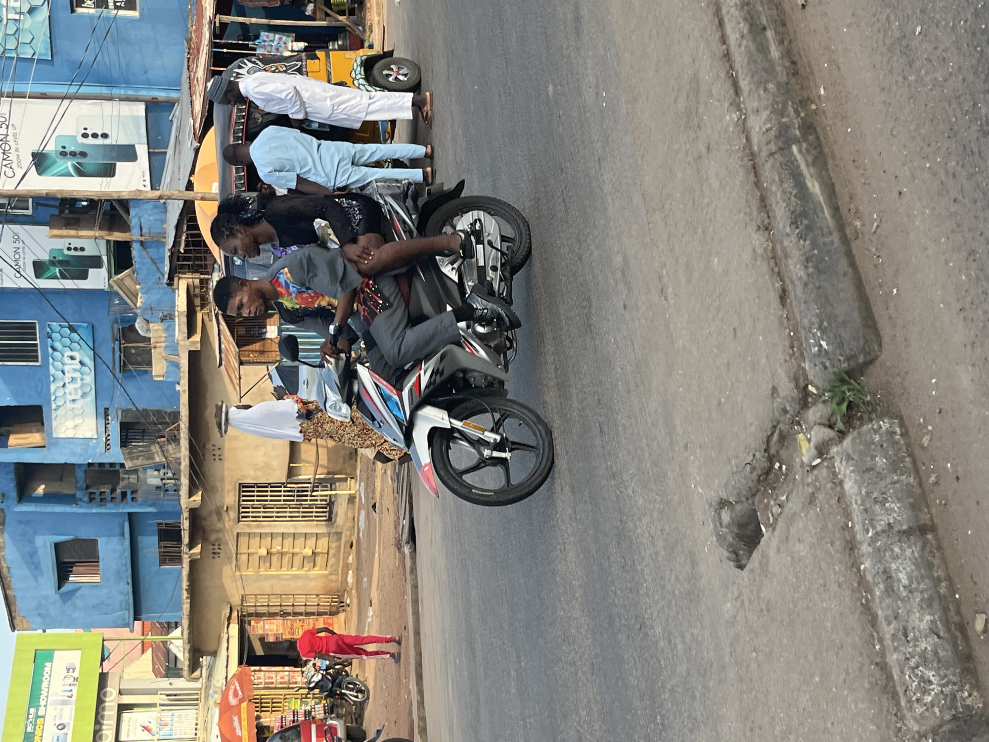 A man and a woman are riding on a motorcycle on a paved road in an urban environment. The man is driving, wearing a patterned shirt and dark pants, while the woman, in a black dress, is riding on the back. They are moving from left to right. In the background, there is a blue building with advertising banners, a smaller tan building with barred windows, and a yellow motorized rickshaw. Several other people are visible in the background, including a man in a red tracksuit standing near a motorcycle, and two men in light-colored clothing near the rickshaw. The scene appears to be a street market or commercial area.
