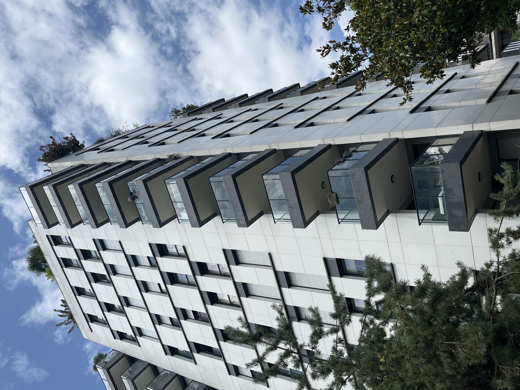 An outdoor, low-angle shot captures a modern, white-tiled apartment building against a blue sky dotted with white clouds. The building features dark gray balconies with glass railings that extend outward from each floor. The balconies are staggered, creating a visually dynamic facade. Trees, one a conifer on the left and another a broad-leafed tree on the right, frame the building. The conifer's branches reach into the lower left portion of the frame. The broad-leafed tree's branches are visible on the right side, partially obscuring the building. Some balconies have potted plants or furniture visible. The building itself has numerous windows and is designed with a contemporary aesthetic. The scene suggests an urban residential environment.
