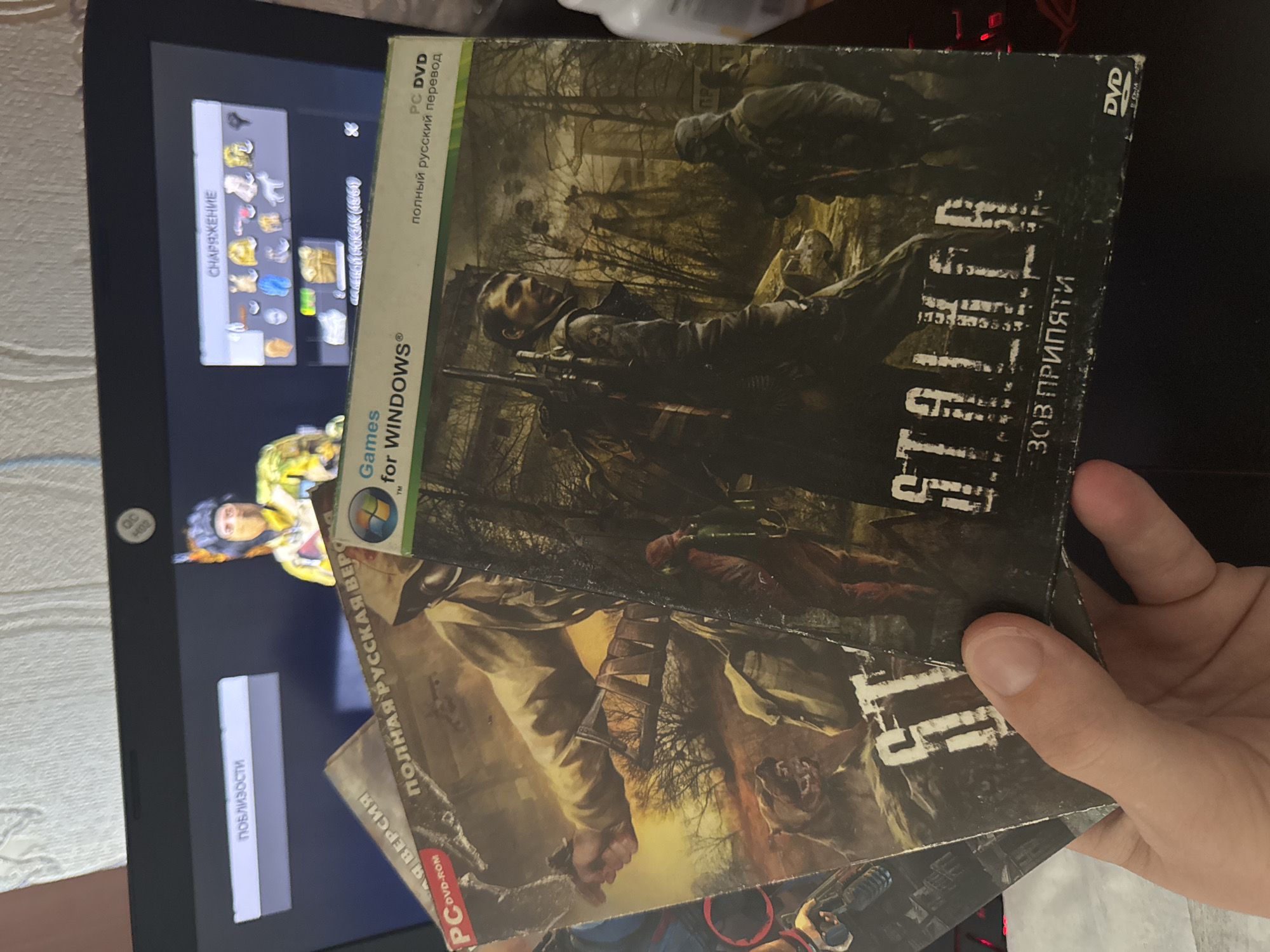 The image shows a person holding two copies of the PC game "S.T.A.L.K.E.R.: Call of Pripyat," both in Russian language editions, in front of a laptop screen. On the screen are in-game menus, including "EQUIPMENT" and "NEARBY." The game is running on Windows, as indicated by the "Games for Windows" logo on the game boxes. The background environment appears to be a desk or workspace. The location is not discernible from the image.