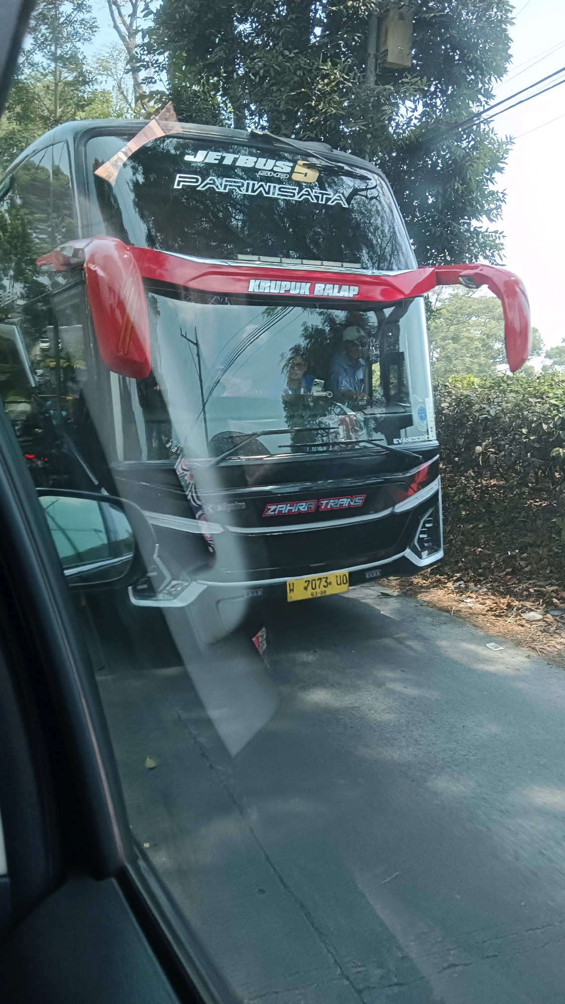 The image shows a front view of a tour bus, likely in Ngargoyoso, Indonesia. The bus is black with red accents and features "JETBUS PARIWISATA" and "KRUPUK BALAP" branding on its exterior. The license plate is visible, reading "W 7073 UO". Inside the bus, two individuals, possibly the driver and a passenger, are visible through the windshield. The bus is on a paved road, with lush green foliage and trees surrounding it, suggesting a rural or semi-rural setting. Sunlight filters through the trees, casting shadows on the road. The image is taken from a low angle, possibly from another vehicle, and reflections on the bus windshield are present.