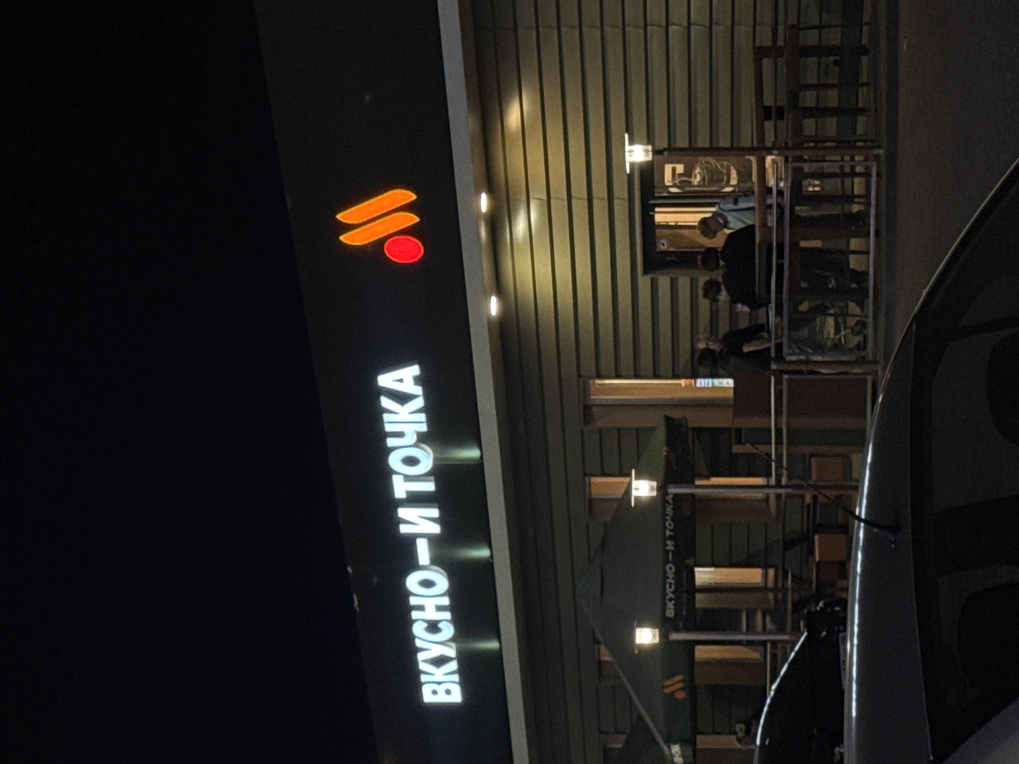 A street view at night depicts a "Vkusno i tochka" restaurant in Izhevsk, Russia. The restaurant's illuminated sign with its logo and name in Cyrillic ("Вкусно — и точка") is prominently visible. Patrons are seen inside the restaurant, while outdoor seating appears to be available. Part of a car is visible in the foreground.