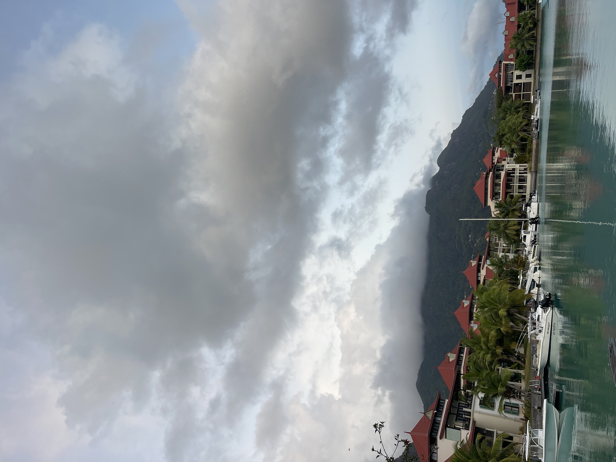 The image shows a waterfront scene in Providence, Seychelles, characterized by a row of buildings with red-tiled roofs situated along a calm body of water. Several boats are docked in the foreground, reflecting on the water's surface. Lush green palm trees are interspersed among the buildings. In the background, a large, mist-shrouded mountain dominates the horizon, with heavy, grey clouds covering the sky. The lighting suggests either early morning or late afternoon, with a subdued, overcast atmosphere. There are no people visible in the image.