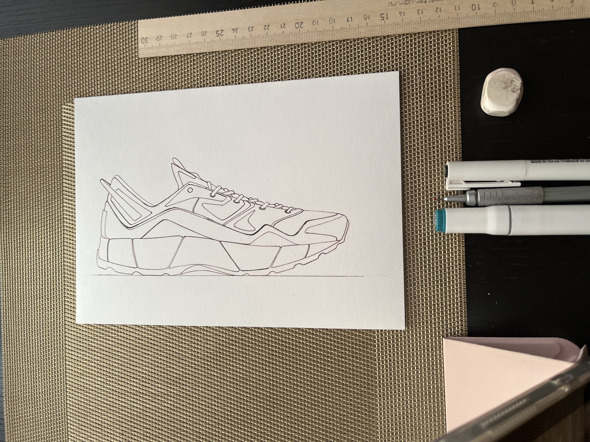 Here's a description of the image:
The image presents an overhead view of a drawing setup on a brown woven mat. The drawing features a detailed line art sketch of a sneaker, rendered in a dark reddish-purple color on white paper.
Arranged near the drawing are art supplies: an eraser, a white marker, a grey technical pen, and another white marker. A ruler with centimeter markings sits above the drawing. A pink object appears in the lower right corner. The background includes a black surface. Based on the ruler's origin information "m.ru" the origin city is Krasnoyarsk, Russia.