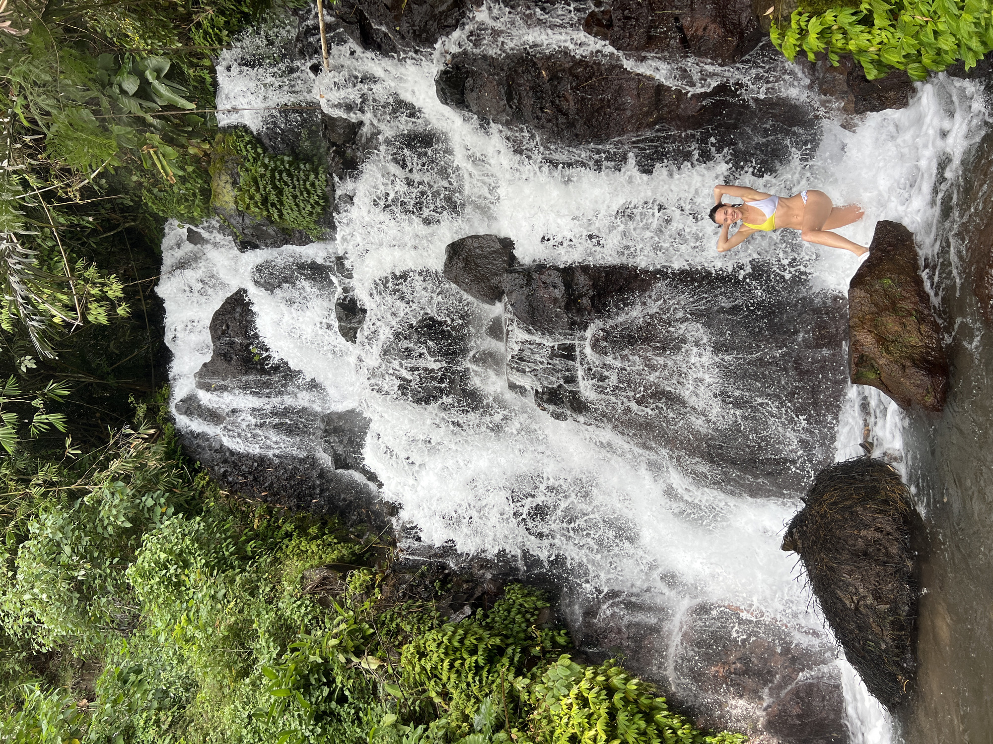 A woman in a yellow and white bikini poses with her hands behind her head in front of a cascading waterfall in Sukasada, Indonesia. The waterfall is surrounded by lush green foliage and dark, wet rocks. The water is turbulent and frothy, creating a misty atmosphere. The woman is standing on a rock in the foreground, with her left leg bent and her right leg extended. She has a smile on her face. The scene appears to be a natural, tropical environment.