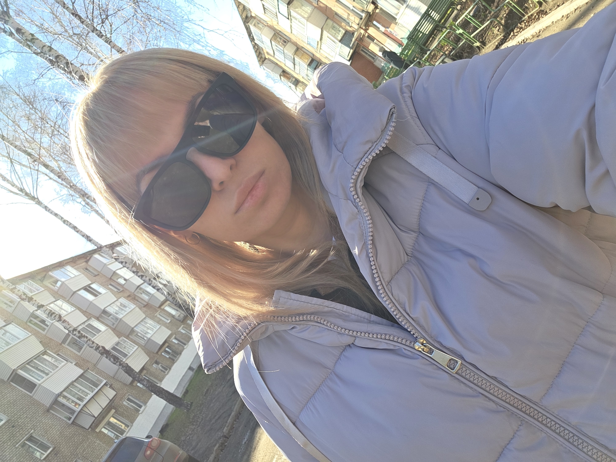 A medium-close-up selfie captures a woman outdoors in Chaikovsky, Russia, on a bright, sunny day. The woman, with blonde hair and bangs, is wearing large dark sunglasses that obscure her eyes, and her lips are slightly pursed in a neutral expression. She is dressed in a light grey or lavender-colored puffer jacket with a prominent silver zipper.
The sunlight is strong, creating bright highlights on her hair and causing some lens flare in the image. In the background, typical urban residential buildings with numerous balconies are visible, suggesting an older architectural style common in post-Soviet countries. Bare birch trees, characteristic of the region and indicating a season other than summer (likely late autumn, winter, or early spring), stand against a clear blue sky. A dark-colored car is partially visible at the bottom left, parked near one of the buildings. The image conveys a casual moment captured in an urban environment. No discernible text is visible in the image.