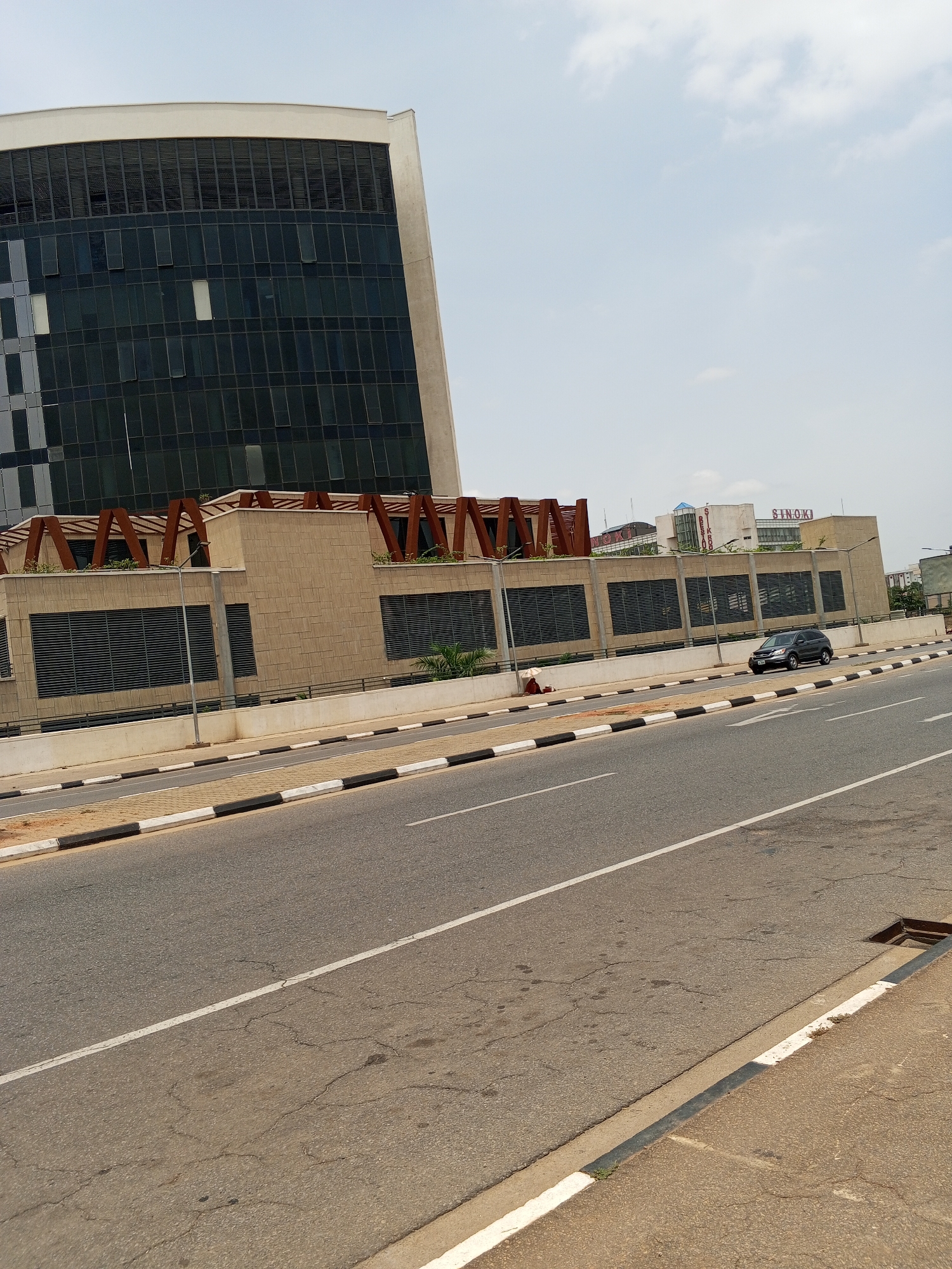 The image depicts a view of a road in Abuja, Nigeria, with several buildings in the background. A modern office building with a curved facade and dark glass windows is to the left. Adjacent to it is a lower, tan-colored structure adorned with linear architectural details and some greenery. On the street, a dark-colored car is visible and a manhole is to the right. The sky is bright, with scattered clouds. The road has a white line marking the lanes and a black and white curb. A sign reading "SINOKI" is visible on a building in the distance.