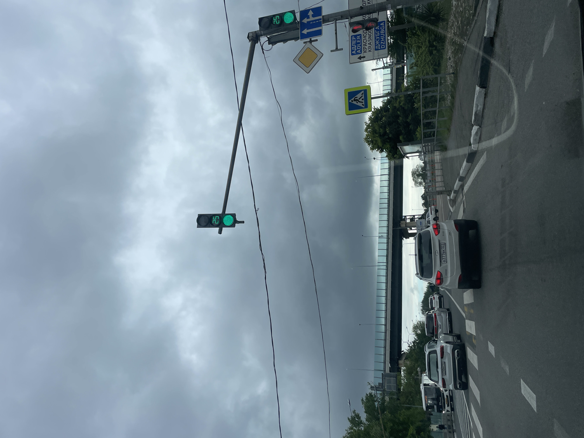 The image depicts a street scene in Sochi, Russia. A row of cars is stopped at a green traffic light. The sky is overcast and cloudy. Overhead directional signs for Adler, Krasnodar, and Dzhubga are visible, as well as a pedestrian crossing sign and a hazard warning sign. A bridge or overpass is visible in the background. The traffic light displays a countdown timer showing 25 seconds.