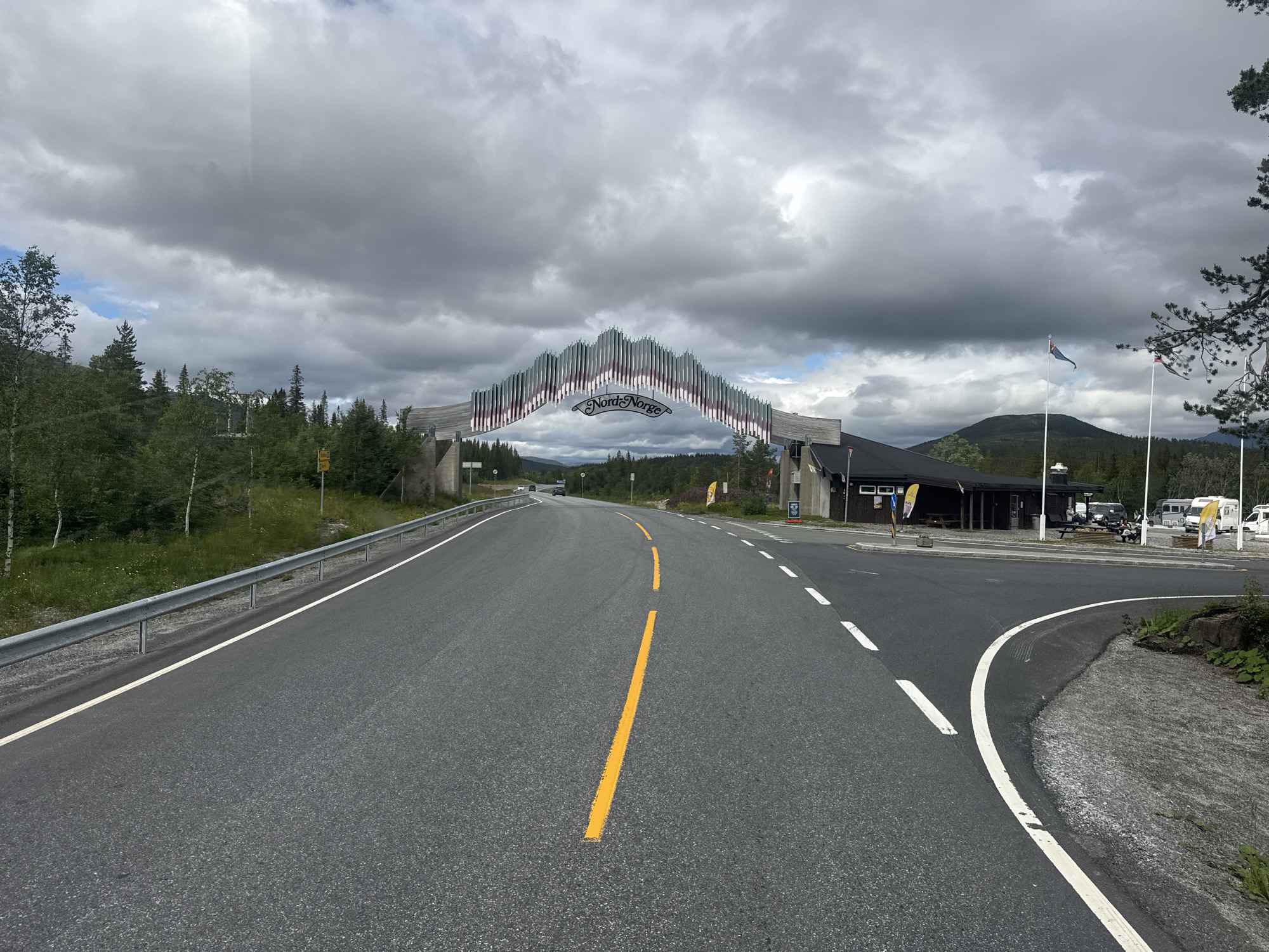 The image shows a road leading towards a large, decorative archway with the text "Nord-Norge" on it, suggesting it marks the entrance to Northern Norway. The archway is situated in a rural or semi-rural setting, with trees and hills visible in the background. There is a building complex to the right of the arch, which appears to be a roadside service area or a small village. Several flags are flying from poles near the building. The road itself is paved and has clear lane markings. The sky is overcast with dark clouds. The location is identified as Smalåsen, Norway. There are no identifiable people in the image.