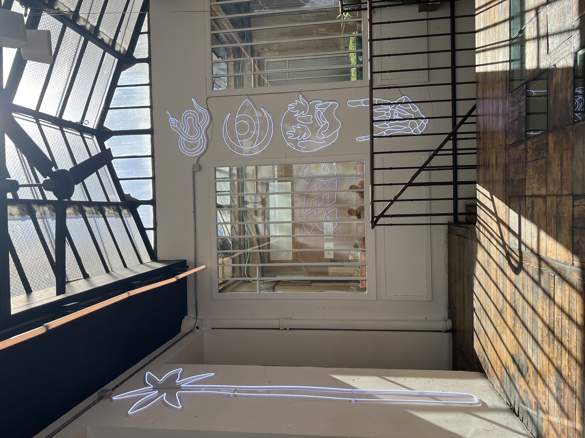 This is an interior shot of a building, likely a cafe or restaurant, in Paris, France. The space features a large, glass-paneled ceiling that allows natural sunlight to stream in, casting diagonal shadows across the wooden floor. Several white neon art installations are visible on the wall. On the left, a neon palm tree shape extends vertically. To the right, a series of neon graphics include a coiled snake, an eye, a face with hands, and a skeletal hand making a peace sign. A black metal railing and staircase are present, suggesting a mezzanine or upper level. The architecture includes white walls, large windows with metal frames, and a black ceiling. Two individuals are partially visible through the windows in the background, seated at a table. The overall aesthetic is modern and artistic.