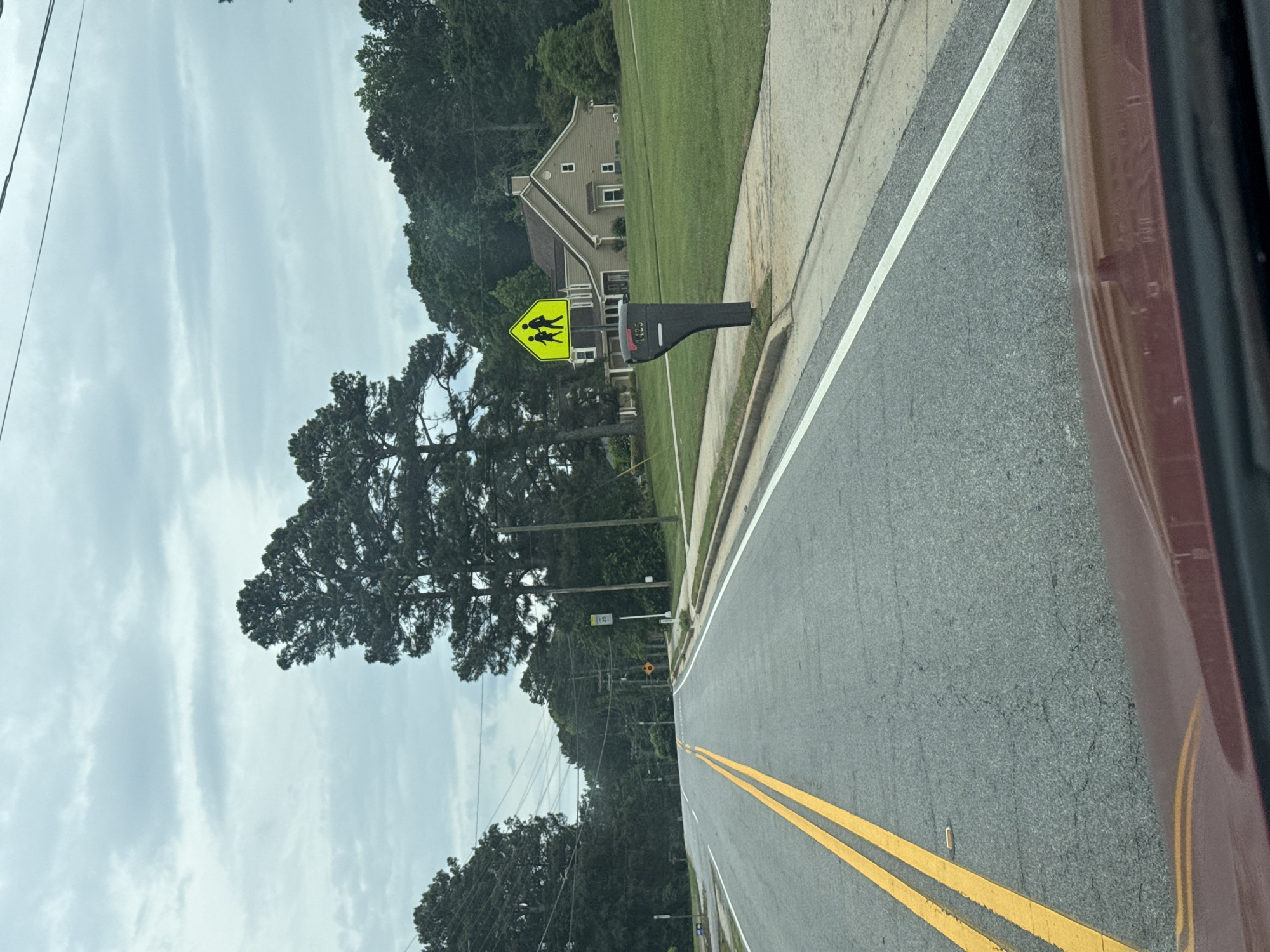 The image depicts a street in Mountain Park, Georgia, USA. The road is paved and marked with double yellow lines. A yellow pedestrian crossing sign is visible on the right side of the road, in front of a residential house. The sky is overcast. Trees line both sides of the road. Part of a car, possibly the vehicle from which the picture was taken, is seen on the right side of the frame.