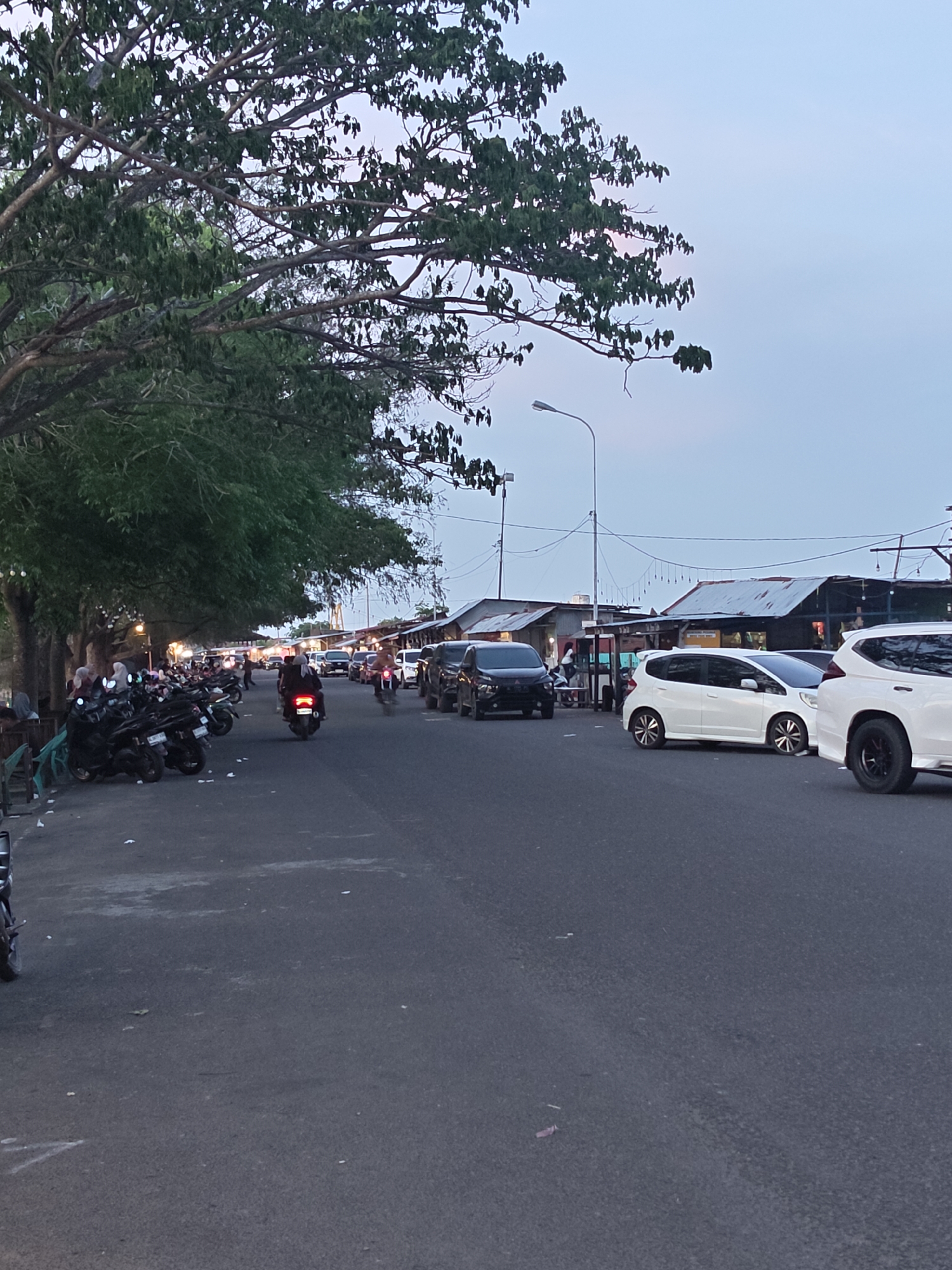 The image shows a street scene in Lhokseumawe, Indonesia. The street is lined with trees on the left side and small shops or stalls on the right. Parked motorcycles are visible along the left side of the street. Cars and motorcycles are driving on the road. The sky is dusky, suggesting either early morning or late evening. The buildings on the right side have simple, tin roofs. Some people are visible standing near the shops.