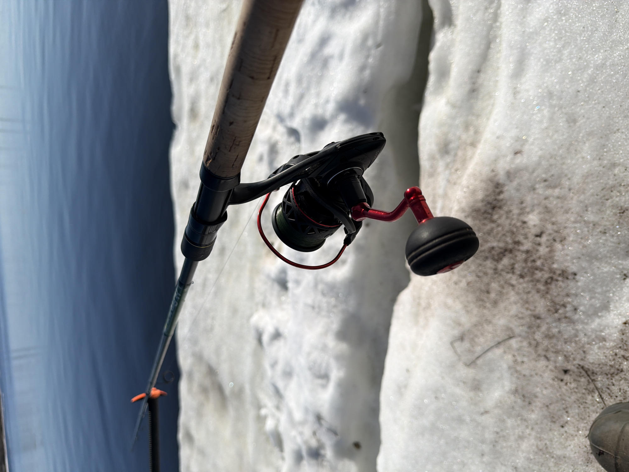 A fishing rod is positioned in front of a snowbank with a body of water in the background, possibly in Chaikovsky, Russia. The rod features a black spinning reel with a red handle. The rod itself is light brown and made of cork. The snowbank is partially dirty, indicating some thawing. The water is a deep blue, and the overcast sky is not visible.