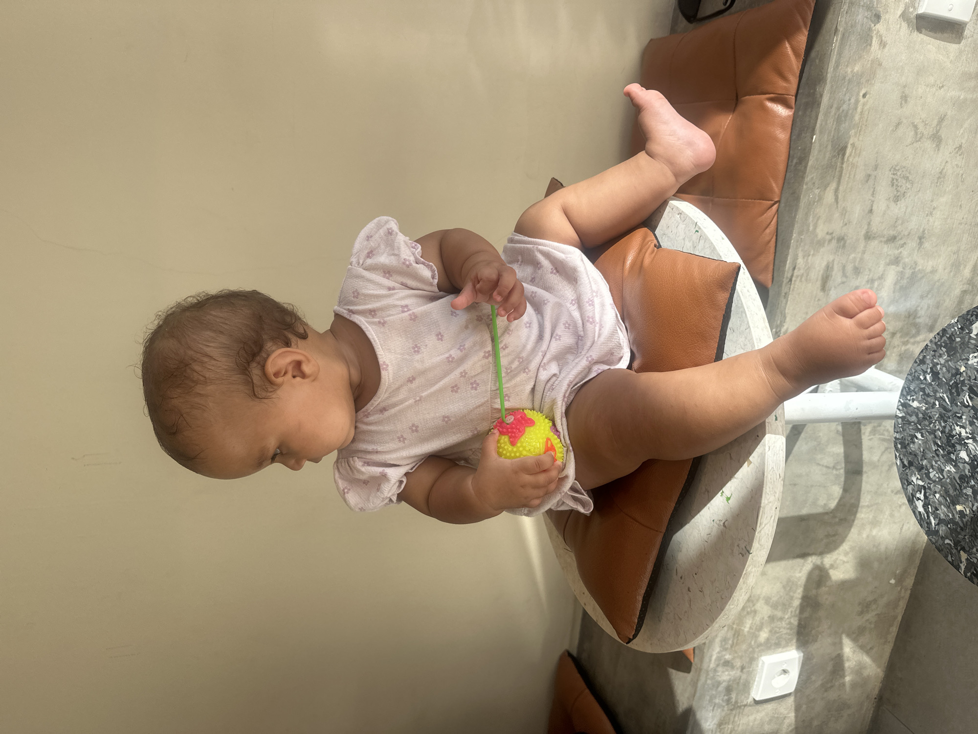 A baby is seated on a chair cushioned with tan leather. The baby wears a light-colored dress with small floral prints and holds a yellow toy with a green handle. The chair sits on a concrete surface in front of a light beige wall. In the background, an electrical outlet is visible. The location is likely in North Kuta, Indonesia.
