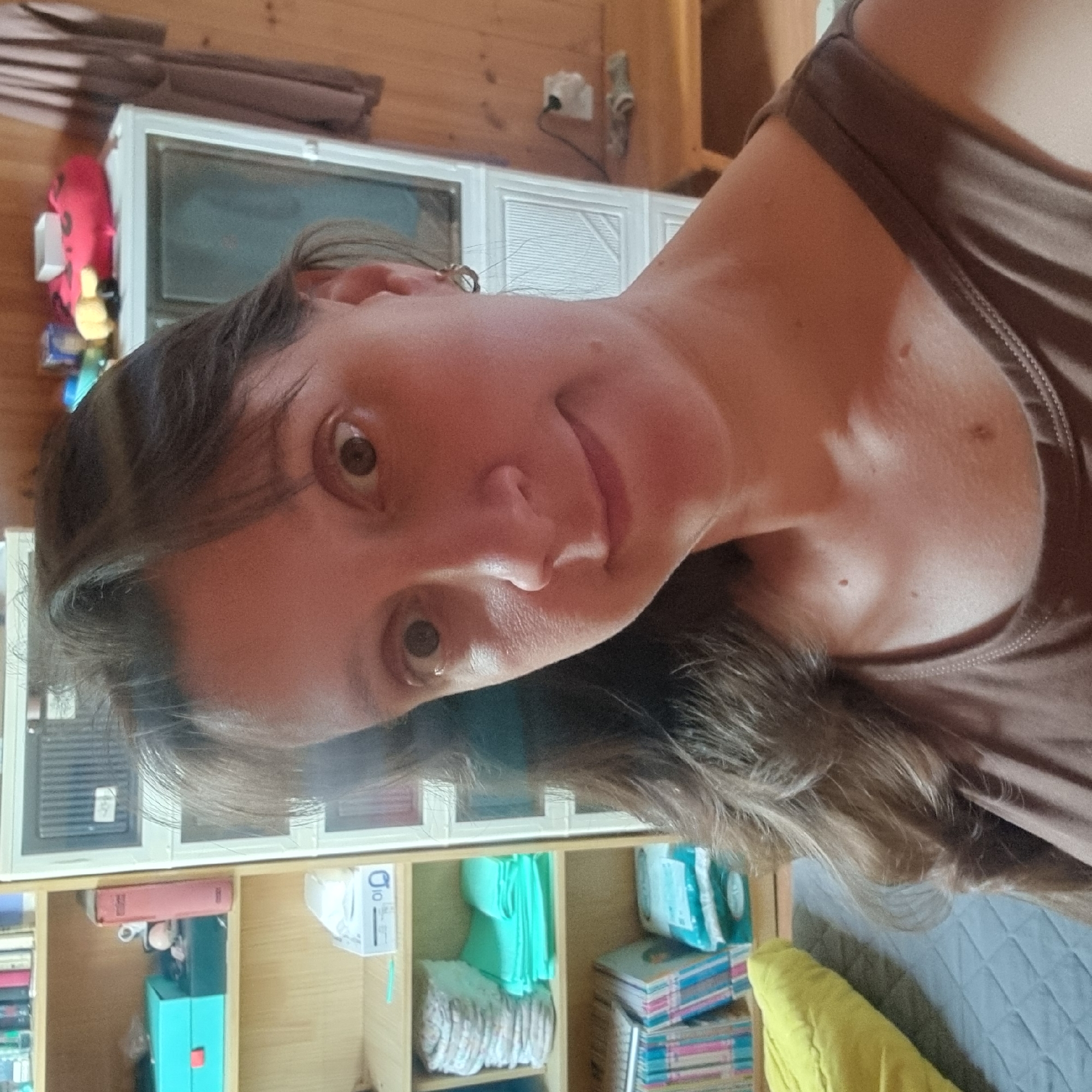 This is a close-up, selfie-style photograph of a woman in Dunnae-myeon, South Korea. The woman has brown hair with bangs and is looking directly at the camera. She is wearing a brown tank top. Her skin tone is fair with some visible moles on her neck and chest. She has light brown eyes and a faint smile.
The background suggests an indoor environment, possibly a room with wooden walls. To the left, a bookshelf is visible, containing various items including what appear to be books, a turquoise box, and stacks of folded cloths or towels. To the right of the woman, a white cabinet or appliance is present. Further in the background, a portion of a wooden dresser or shelf is visible with a black electrical outlet and some items placed on it. A grey quilted mat is in the lower left of the frame with a yellow pillow on it. The overall lighting appears natural and somewhat diffused.
