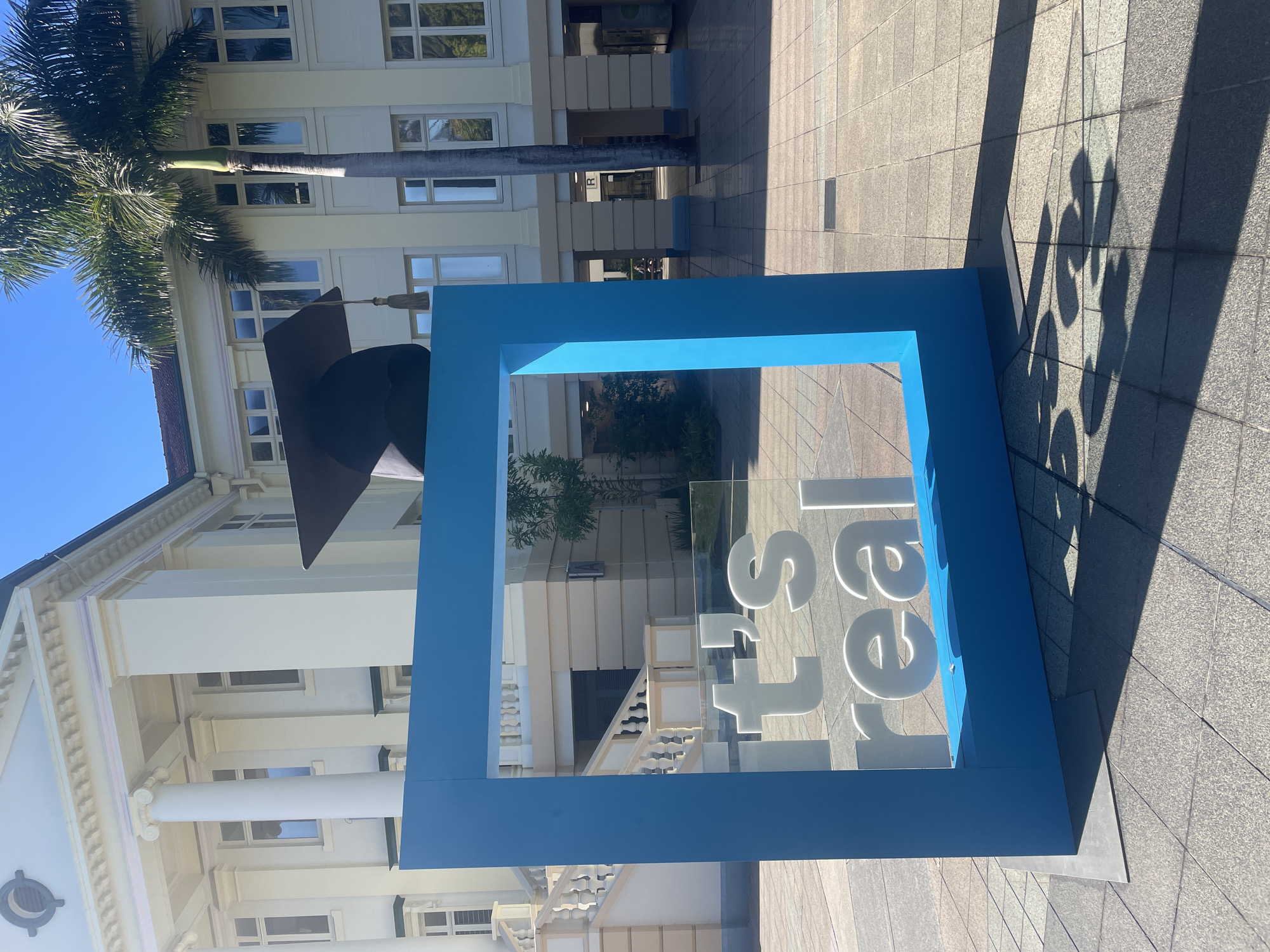 The image captures a sunlit outdoor scene in Kelvin Grove, Australia, with a classic building featuring columns and multiple windows in the background. In the foreground, a bright blue rectangular frame stands prominently, displaying white text on a transparent surface that reads "it's real". Behind the frame, a large mortarboard, a symbol of graduation, is positioned. A tall palm tree adds a tropical element to the environment. The ground is paved with square tiles, and the scene is characterized by strong shadows cast by the objects and the sun. No people are visible in the image.