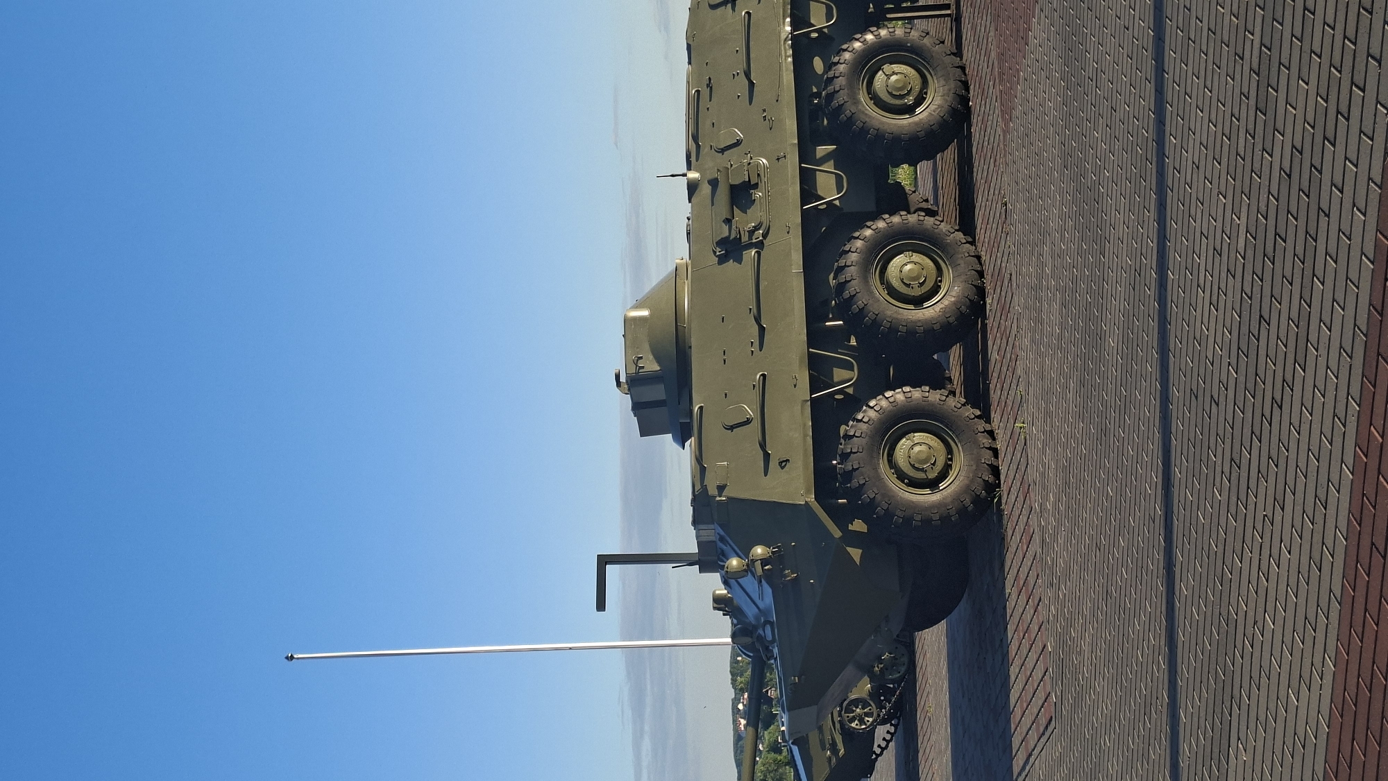 The image shows two military vehicles parked on a brick-paved surface in front of a clear blue sky. The vehicle in the foreground is a large, olive-drab armored personnel carrier with eight wheels. It has a distinctive turret on top and various hatches and armored plating. To the left and slightly behind it, a portion of another military vehicle, possibly a tank with tracks and a barrel, is visible. A tall, thin white pole stands to the left of the vehicles, casting a shadow. In the background, a line of trees and buildings can be seen under the horizon. The scene appears to be an outdoor exhibit or memorial, possibly in the city of Smolevichi, Belarus, given the context. There are no people visible in the image.
