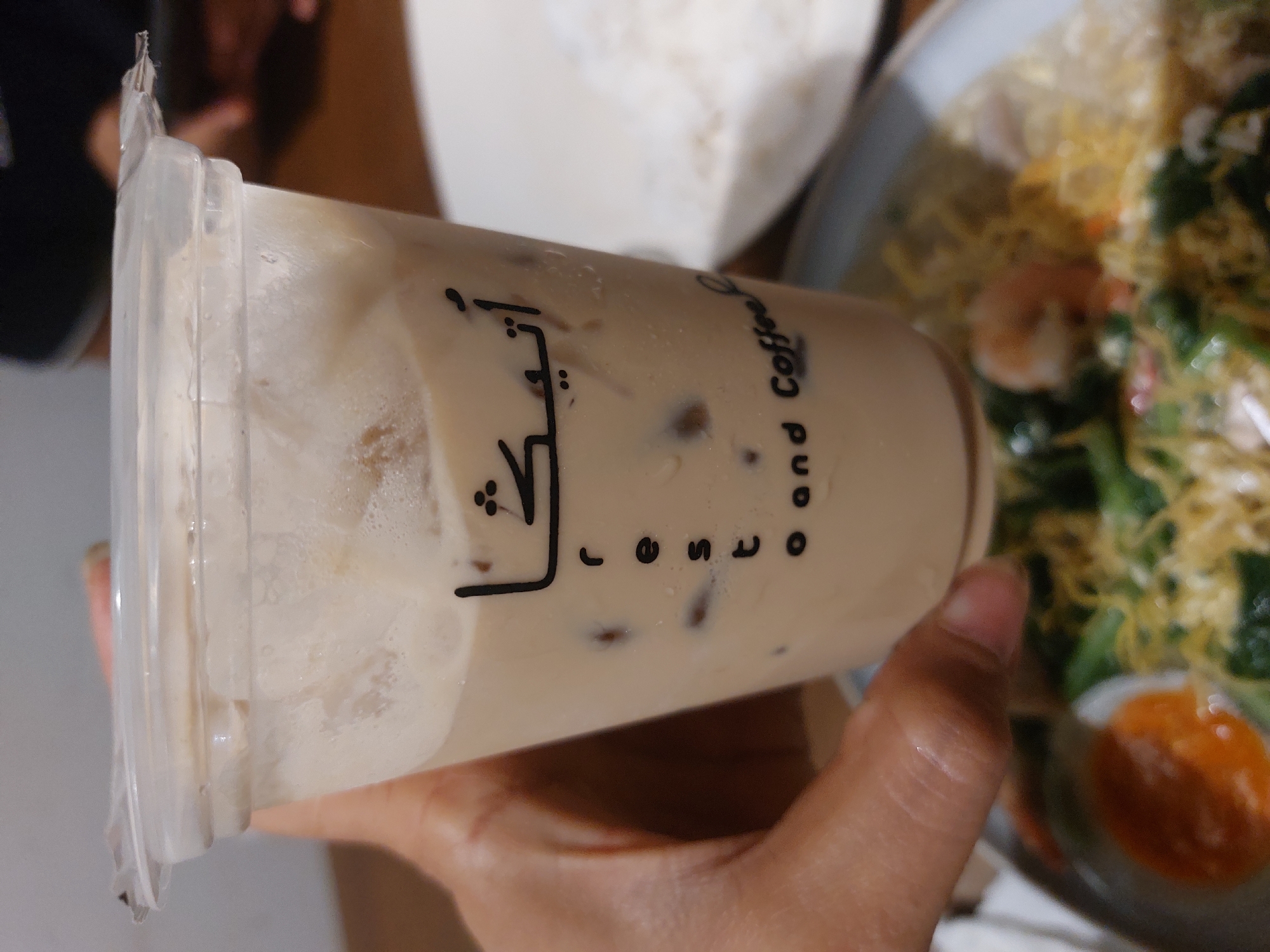 A person's hand holds a clear plastic cup filled with iced coffee. The cup has black Arabic lettering on it, which reads "أتيكش" (Atiqah), and below it, "resto and Coffee". The coffee is a light brown color with milk and ice. In the background, a plate of food with noodles, vegetables, and shrimp is visible, along with a plate of white rice. The setting appears to be a restaurant in Nunukan, Indonesia.