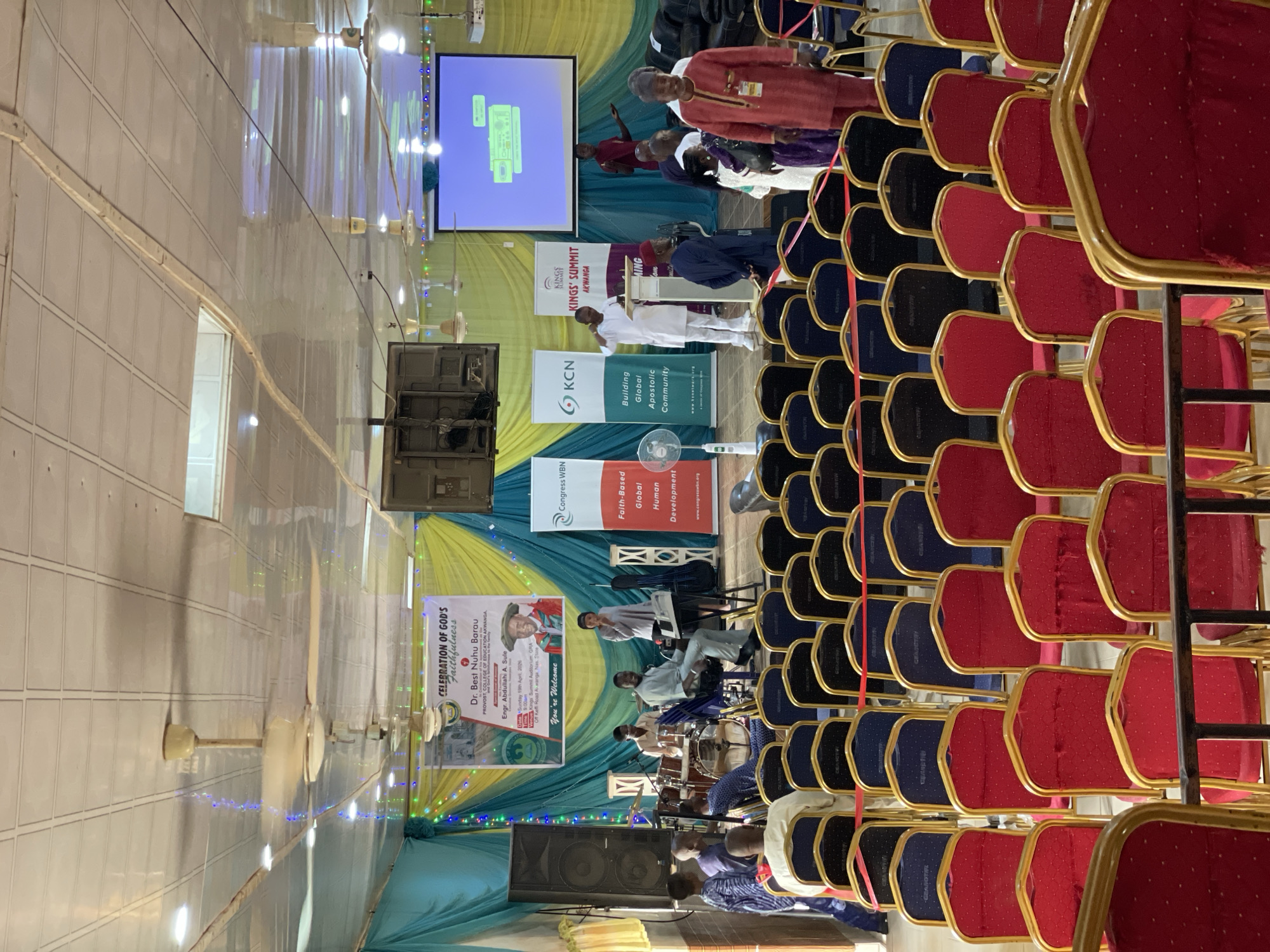 The image shows a conference or event taking place in Akwanga, Nigeria, as indicated by a sign that reads "Kings' Summit Akwanga". Rows of red and blue chairs are arranged in the foreground, suggesting a seating arrangement for attendees. At the front, there's a stage or platform with various speakers and a band. Banners and posters are visible in the background, promoting the event and mentioning "Celebration of God's Faithfulness" for Dr. Best Nuhu Barau, the Provost of the College of Education in Akwanga, along with Engr. Abdullahi A. Sule. The event venue is noted as Kings Summit Auditorium GRA, off Keffi Road. A screen displays a presentation, and the room is lit with ceiling lights and decorated with draped fabric in shades of yellow, green, and blue. A few ceiling fans are also visible. People are seated and participating in the event.