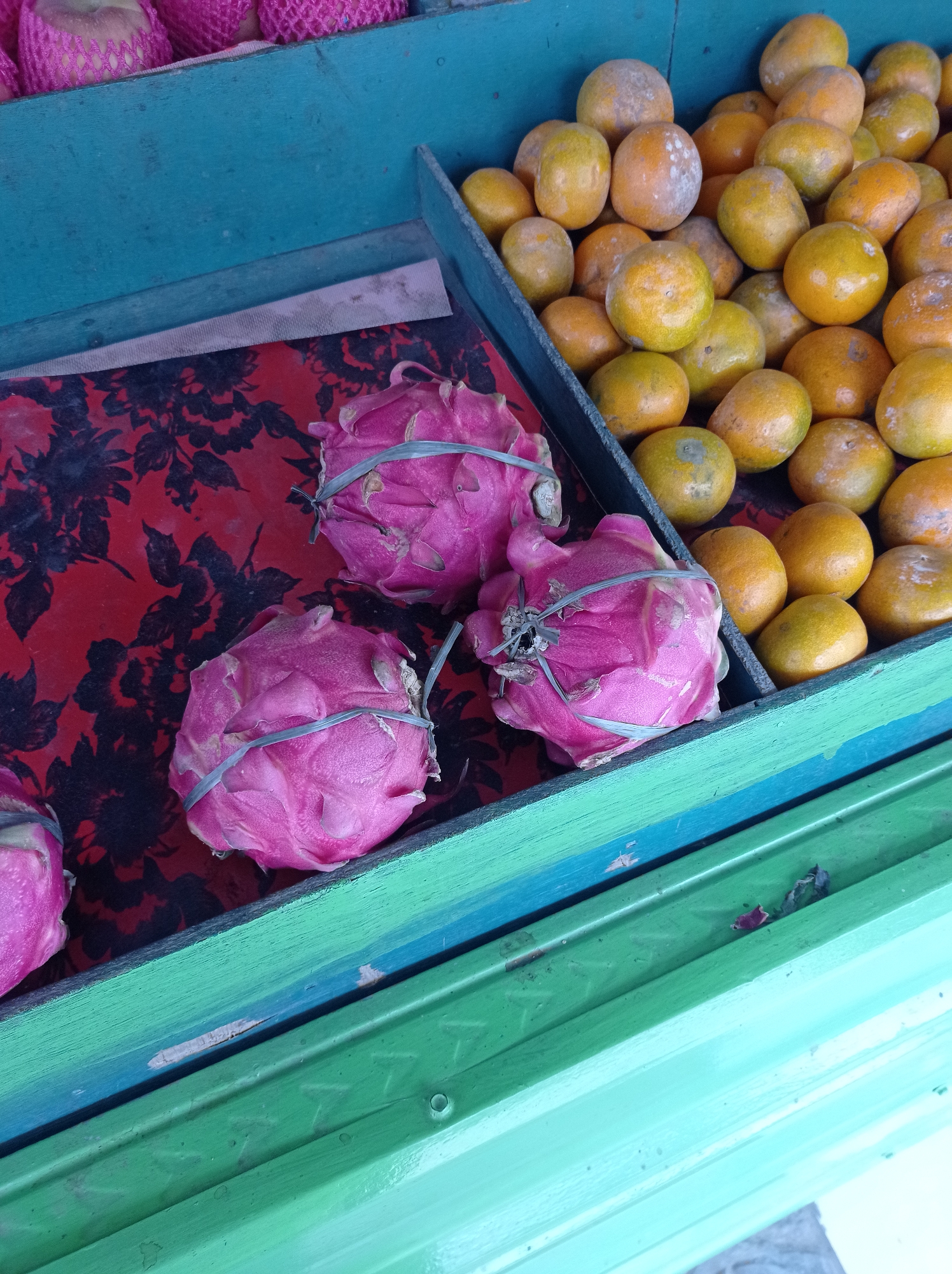 A close-up, top-down view shows a stall displaying fresh fruits. In the foreground, three bright pink dragon fruits are arranged on a red cloth with a black floral pattern. Each dragon fruit is secured with a thin, gray rubber band. To the right, a wooden crate filled with small, round, yellow-orange fruits, likely mandarins, is visible. The fruits are a vibrant color and appear to have a slightly dusty or weathered texture. The stall itself is constructed of painted wood, with the crates painted in contrasting shades of teal and green. The image appears to be taken in Kramat, Indonesia, capturing a slice of local market life.