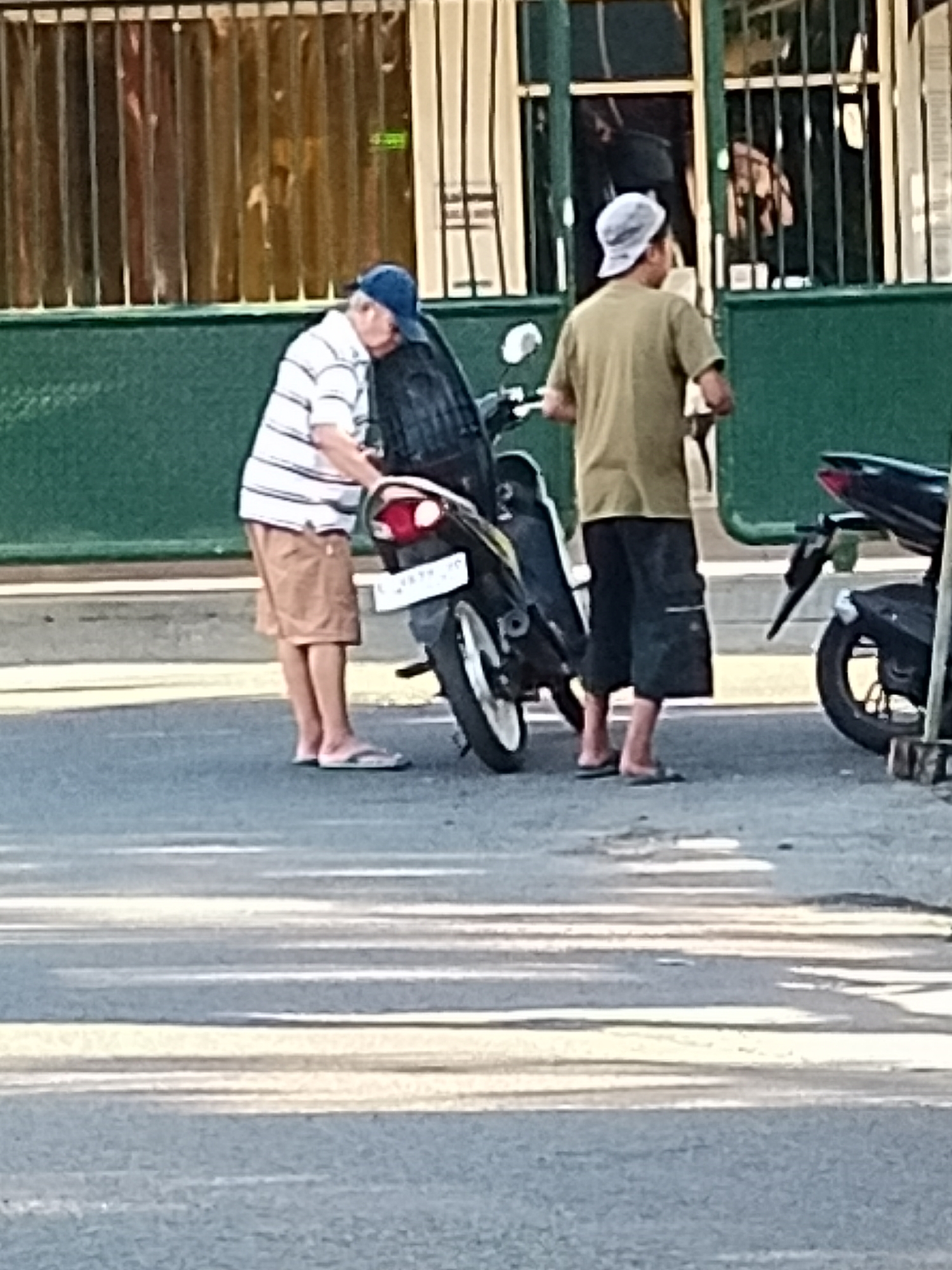 The image depicts two men standing near motorcycles on an asphalt road in Surabaya, Indonesia. The man on the left, wearing a striped t-shirt, blue cap, and tan shorts, is leaning towards a black motorcycle. The man on the right, wearing a tan t-shirt and dark shorts, is facing away from the viewer and standing beside another motorcycle. The background features a building with vertical blinds and green metal gates. The overall scene suggests an urban street environment, possibly a roadside or parking area.