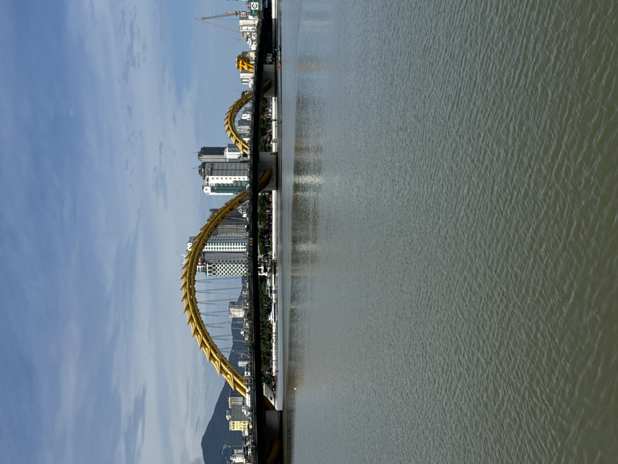 The image shows a view of the Dragon Bridge in Da Nang, Vietnam, spanning across the Han River. The bridge, a prominent landmark, features a distinctive golden dragon design arching over the roadway. In the background, a city skyline rises with various buildings, including modern high-rises and some older structures. A green, treed embankment runs along the river. The water of the Han River fills the foreground and reflects the sky and bridge. The sky is partly cloudy with a bright, hazy atmosphere. There are several boats on the river, including a few small vessels with people in them near the bridge. Construction cranes are visible in the distance, suggesting ongoing development. The overall scene depicts a daytime urban landscape with a focus on the iconic bridge and its surrounding environment.