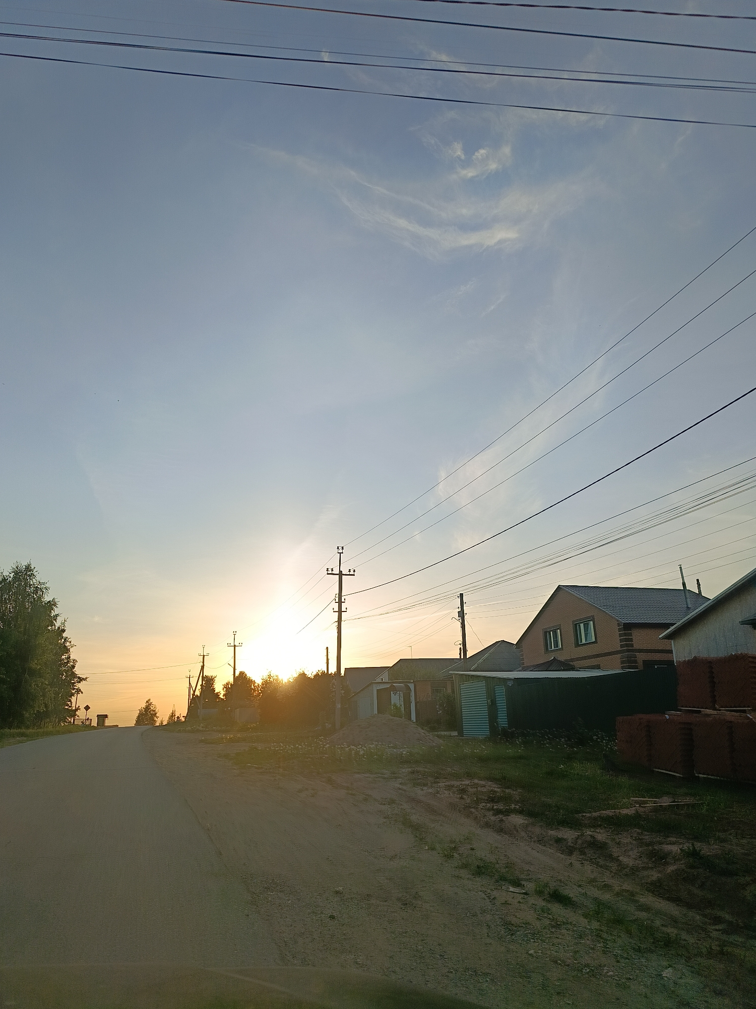 The image depicts a street scene in the city of Chaikovsky, Russia. A paved road stretches into the distance where the sun is setting, creating a bright glare. Overhead, power lines crisscross the sky. Houses are situated on either side of the road, with brick and siding visible on their exteriors. A stack of what appears to be brown bricks is located near one of the houses. Vegetation, including grass and trees, flanks the road. The overall atmosphere suggests a quiet, residential area during the late afternoon or early evening.
